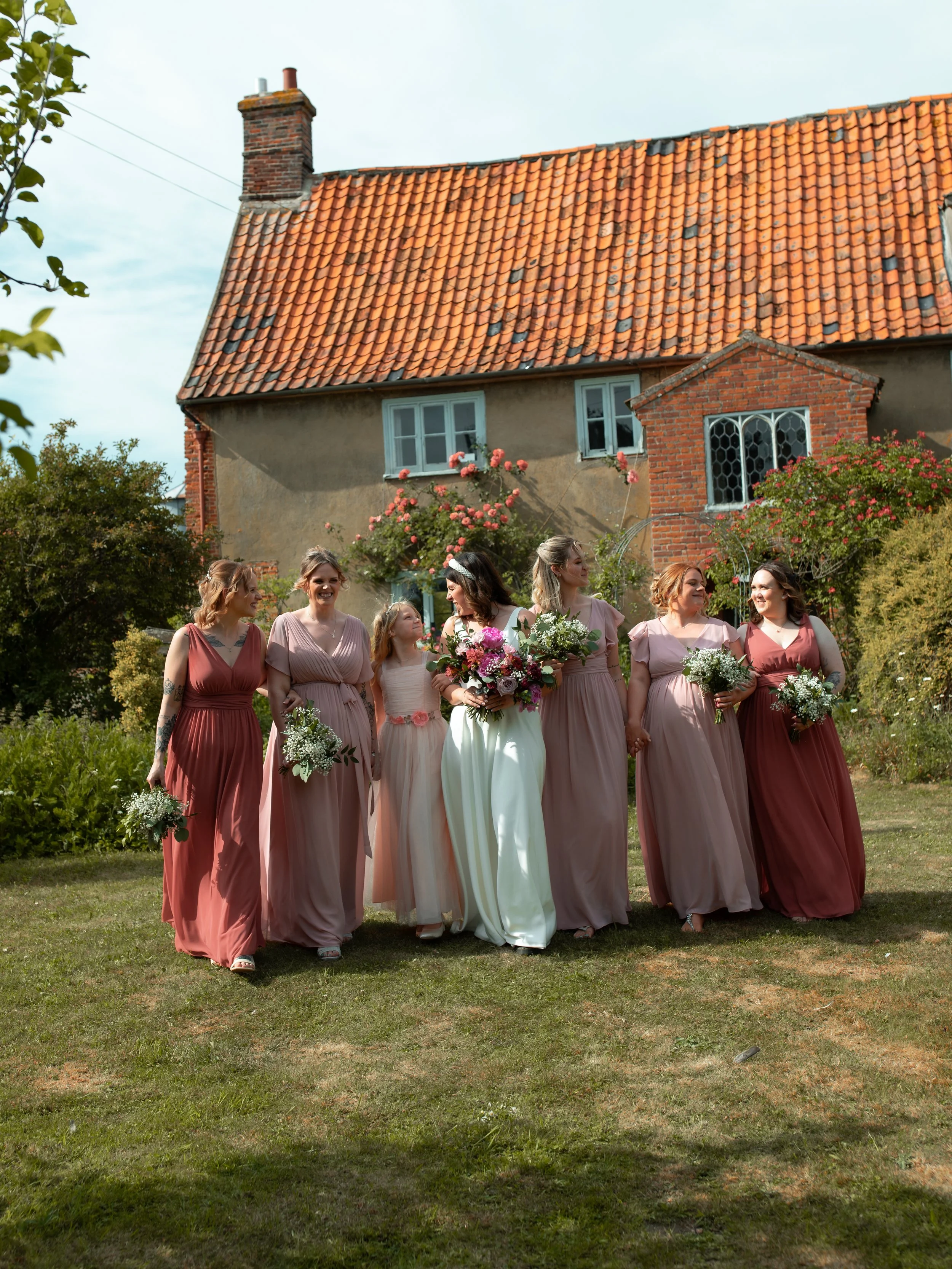 A woman in a white dress holding a Bouquet of flowers, surrounded by women in pink dresses, walking on a grassy lawn in front of a house with a red-tiled roof and a brick chimney.