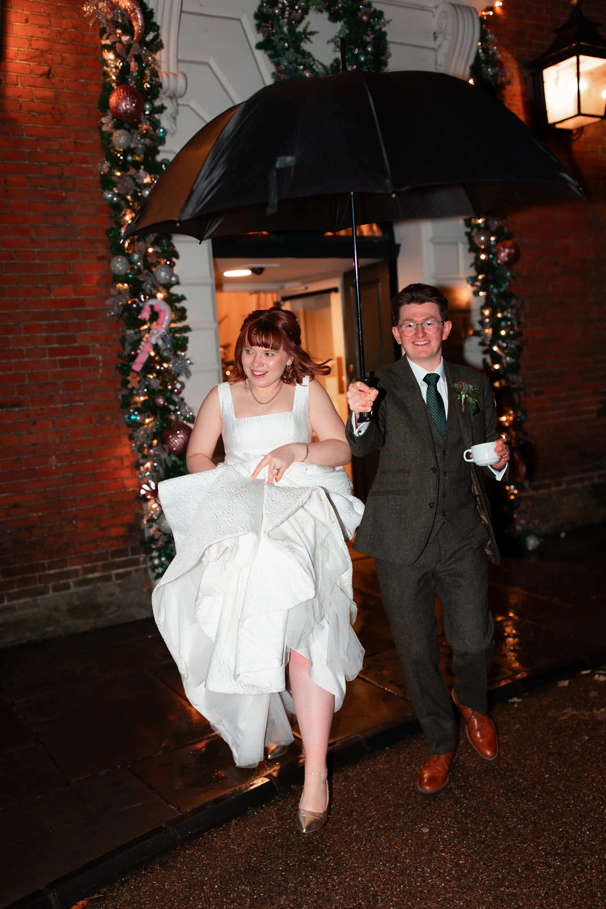 A couple walking under an umbrella outside during a rainy night, with Christmas decorations and lights in the background, and the woman in a white wedding dress.