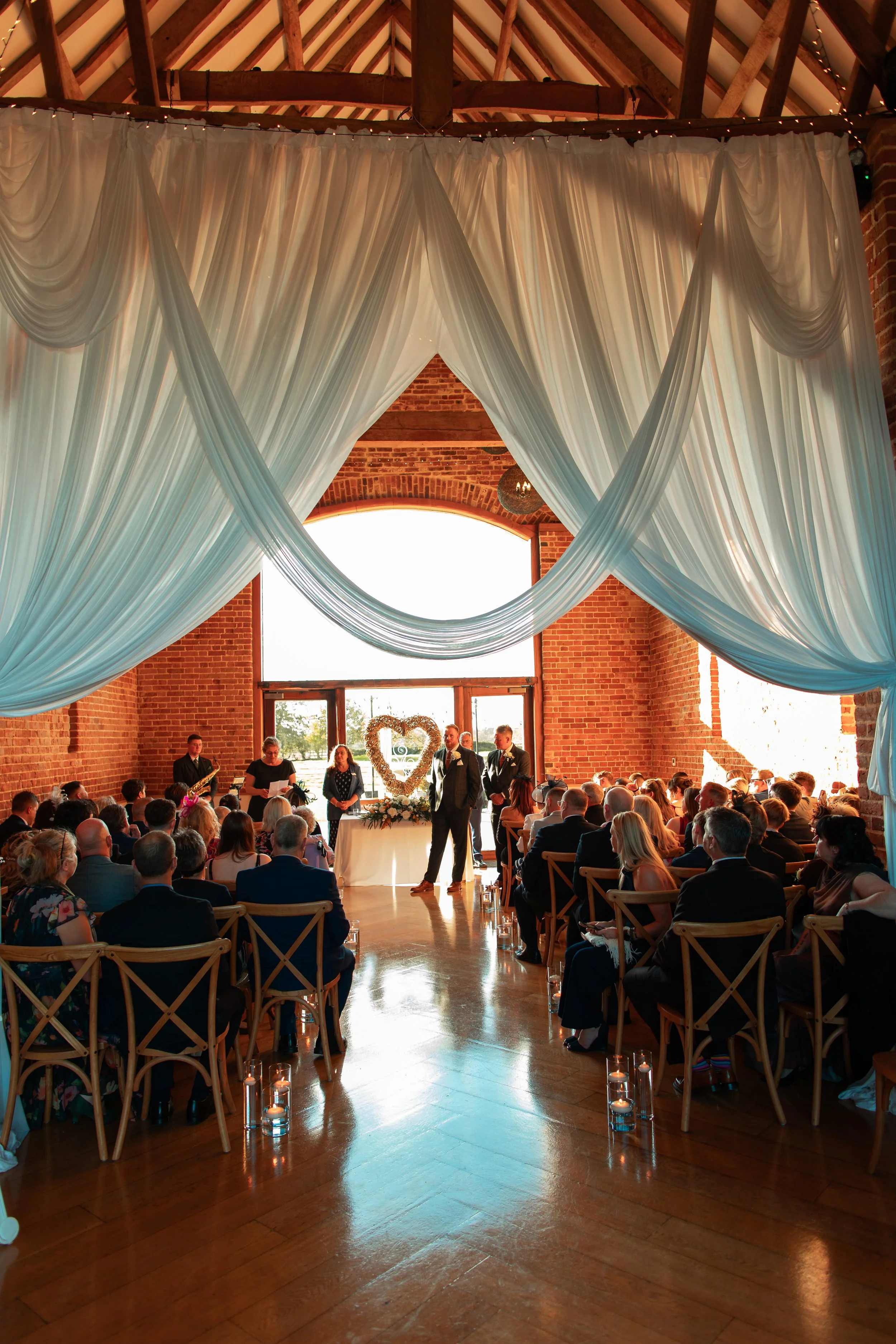 A wedding ceremony inside a rustic venue with exposed brick walls and large arched windows. The officiant is reading, and the couple is standing in front of a heart-shaped floral arrangement. Guests are seated on wooden chairs, and hanging white drap