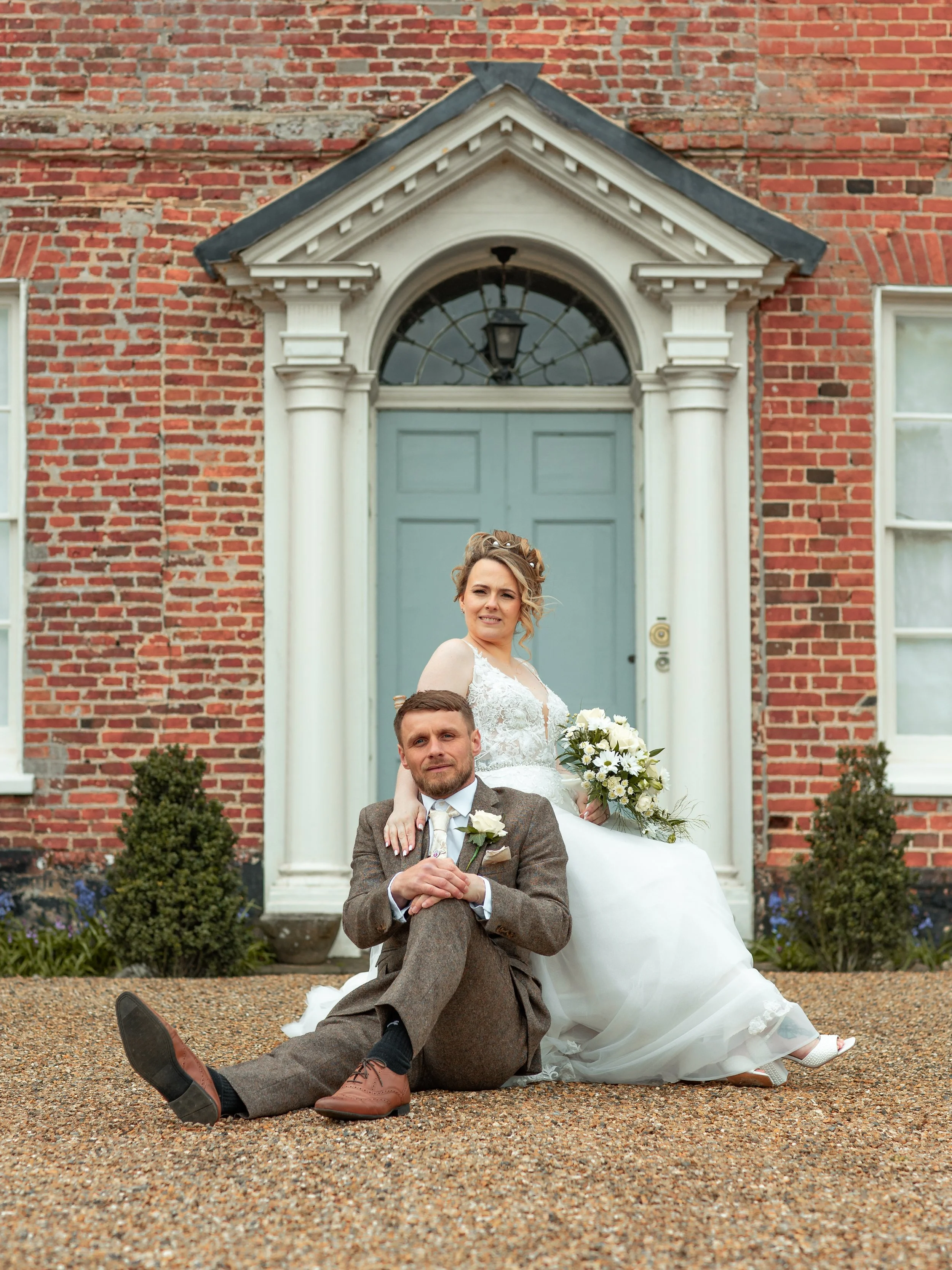 A newlywed couple sitting in front of a brick house with a blue door. The bride is standing behind the groom, holding a bouquet of white flowers, wearing a white wedding dress. The groom is seated on the ground with crossed legs, dressed in a brown t
