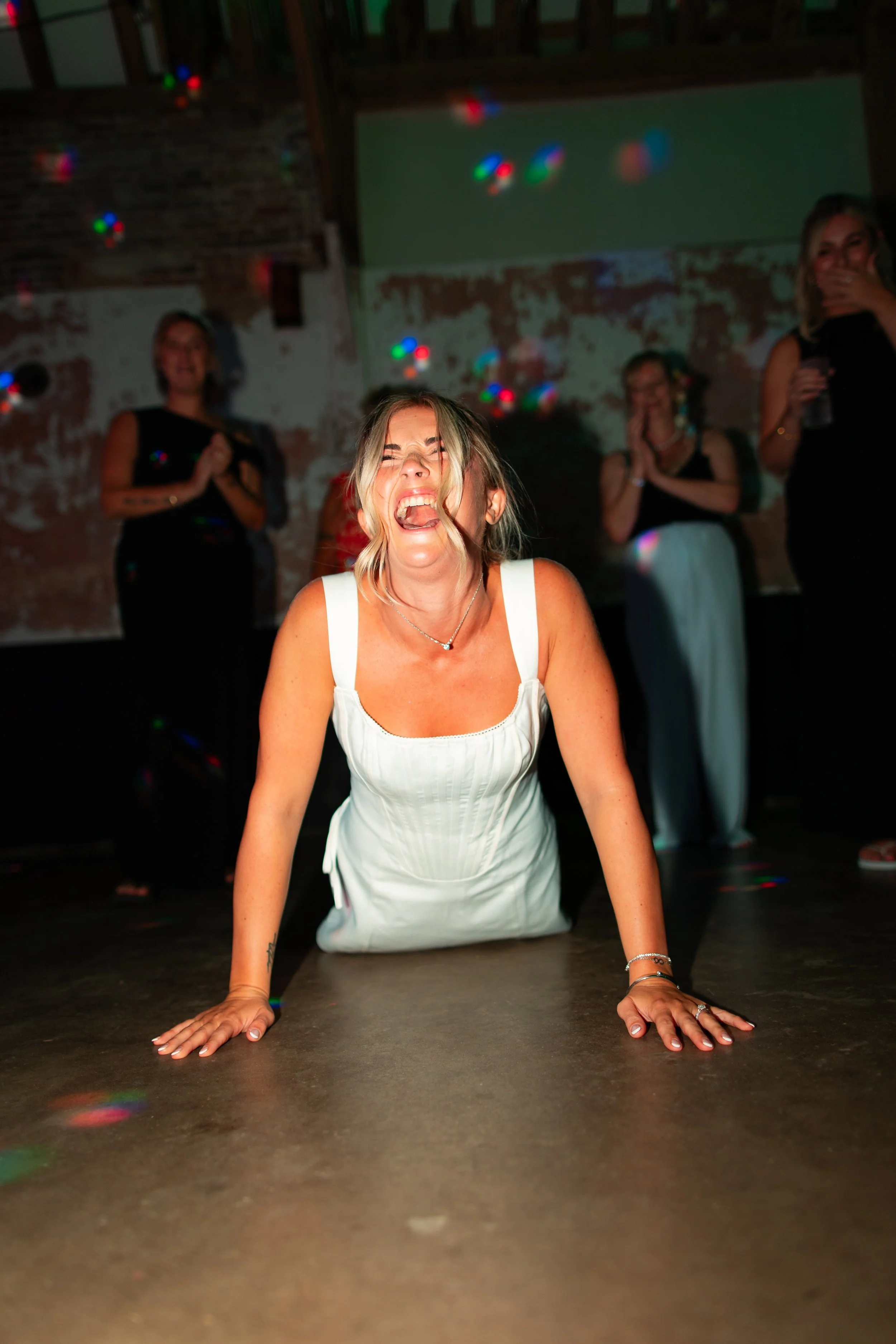 A woman in a white dress on her hands and knees, laughing and enjoying herself at a party with women in black dresses in the background.