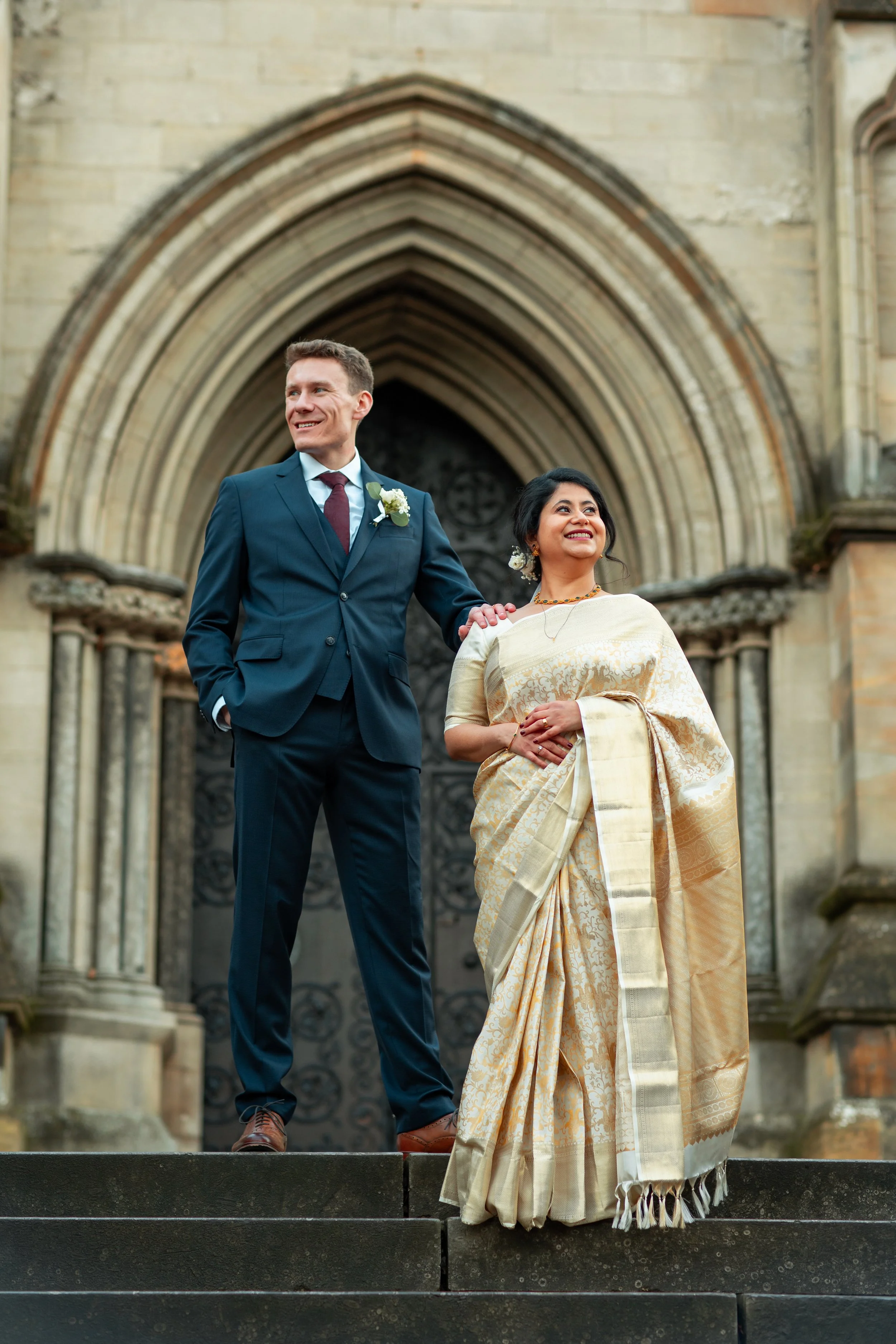 A couple dressed in wedding attire standing outside a historic stone building with gothic architecture, possibly a church or cathedral.