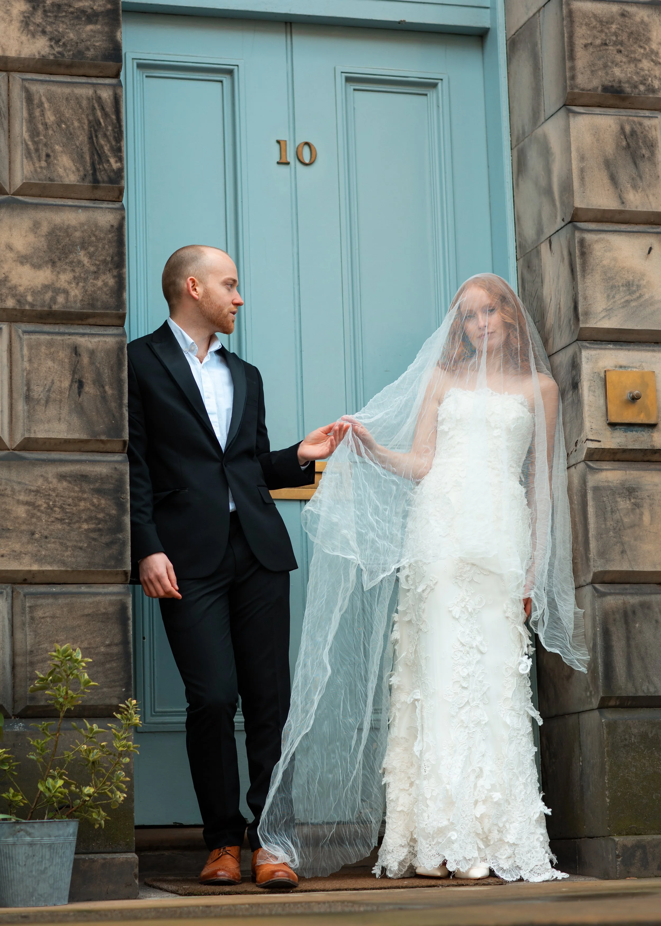 A bride and groom standing outside a building with a light blue door, holding hands, with the bride in a white wedding dress with a veil and the groom in a black suit and brown shoes.
