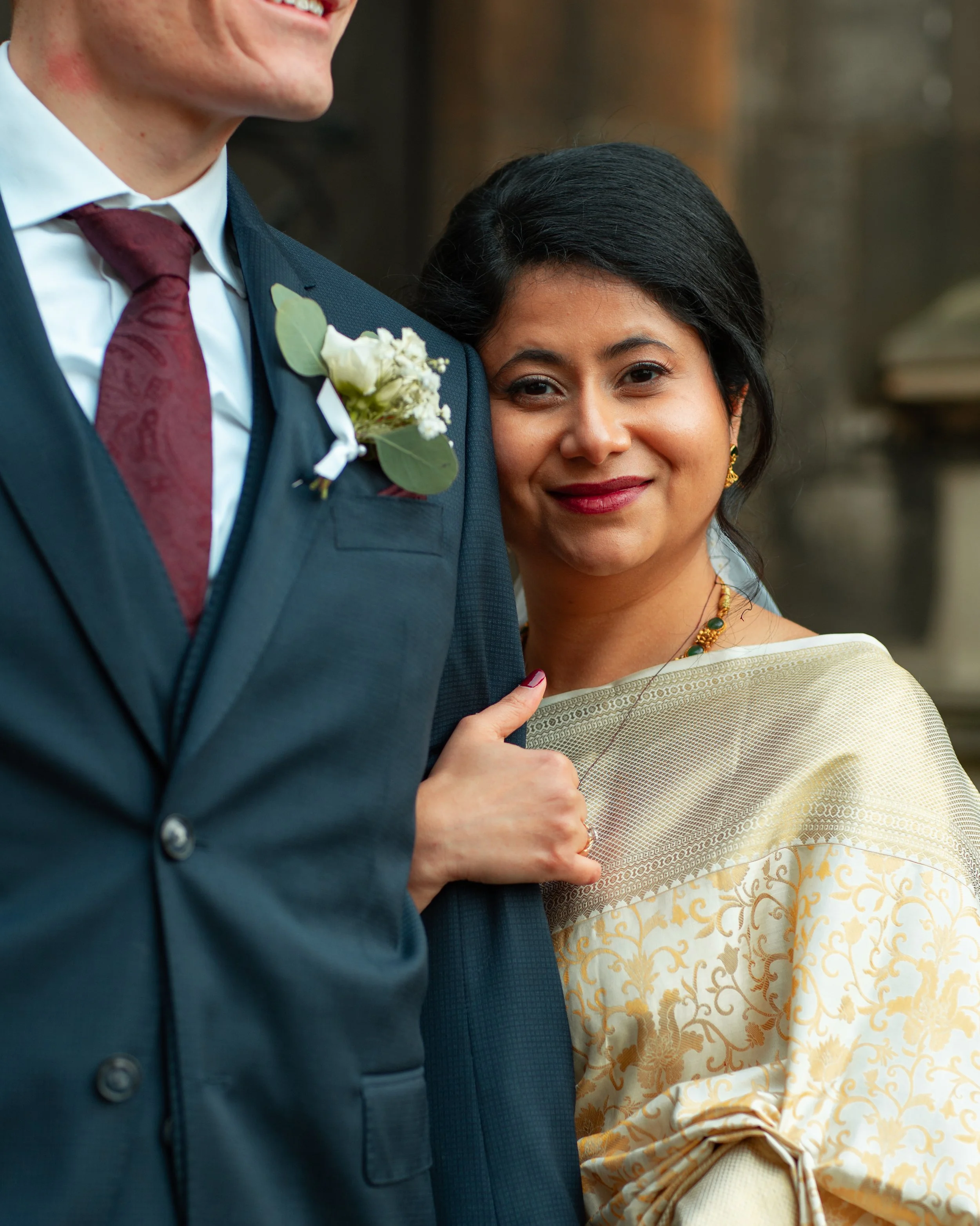 A woman wearing a cream-colored traditional outfit with gold patterns, holding onto a man's suit jacket and smiling.