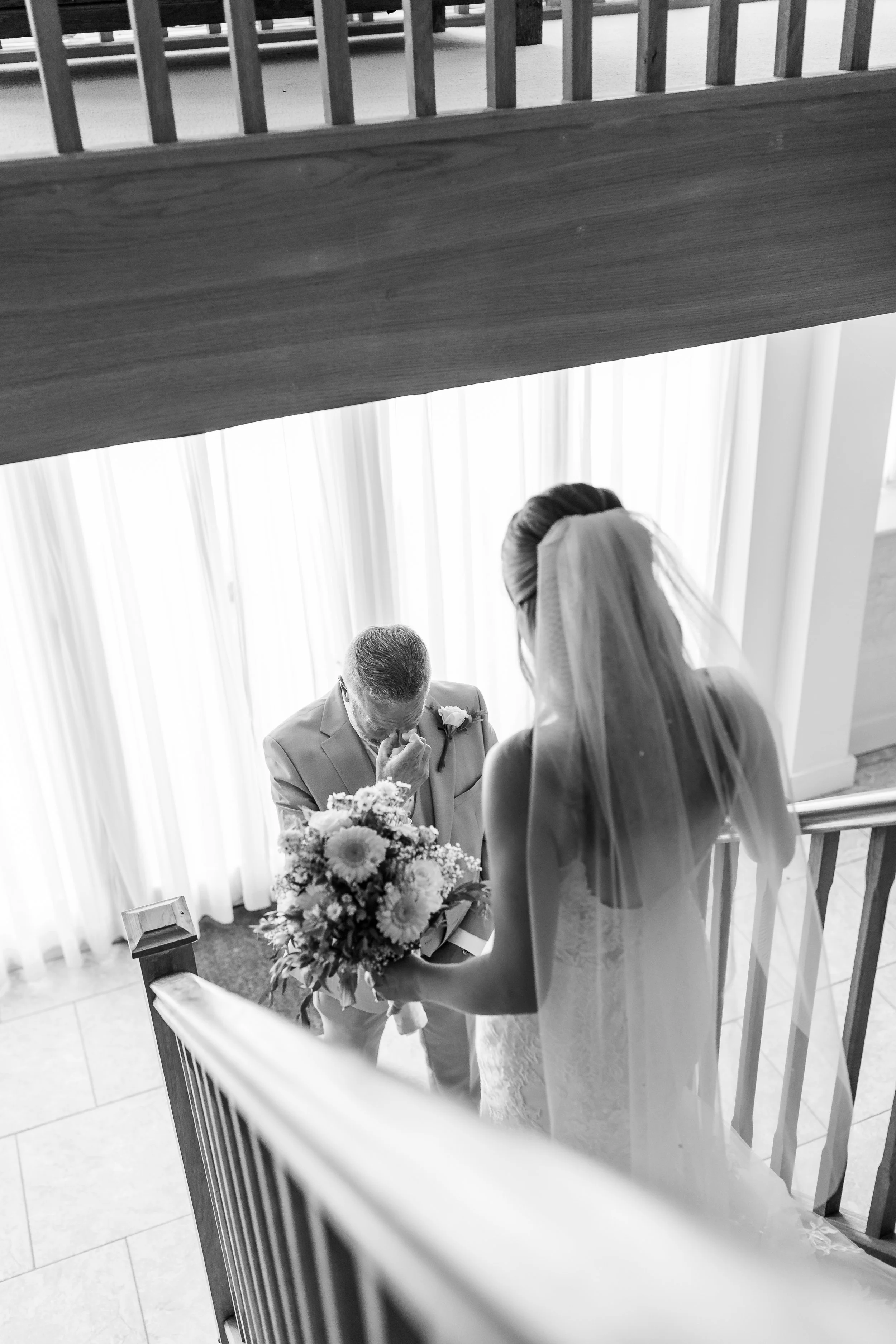 A bride holding a bouquet standing on a staircase, crying as she looks at an older man in a suit, likely her father, who is wiping his tears.