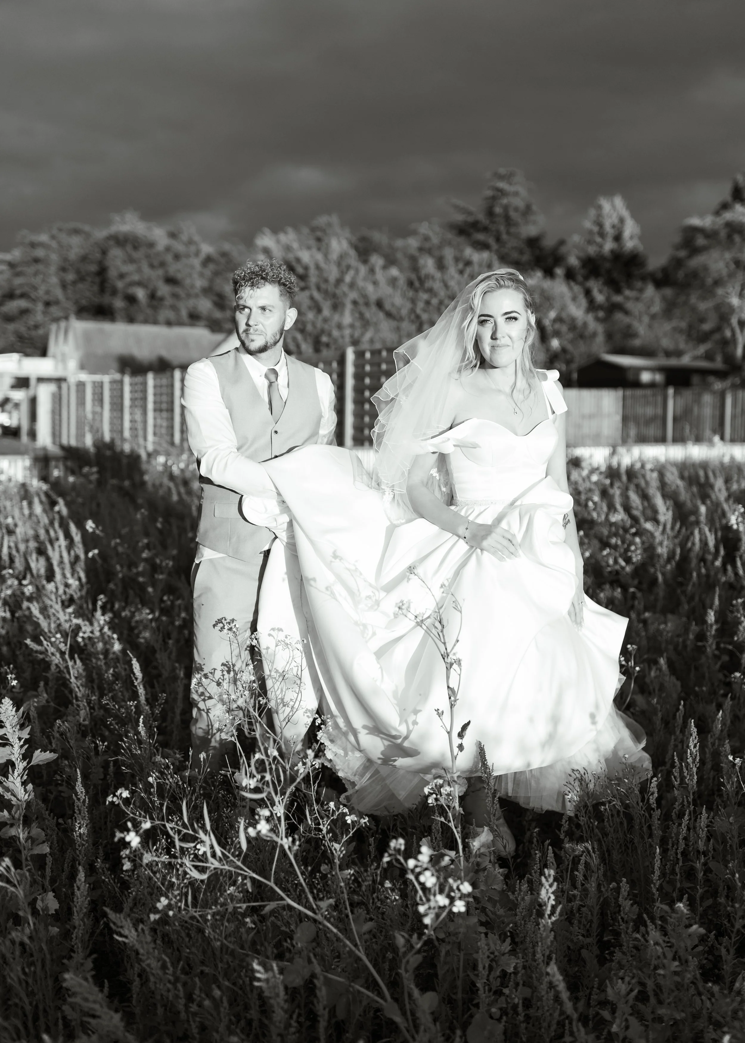 A black and white photo of a bride in a wedding dress and veil being carried by a groom in a vest and dress shirt, standing in a field of tall grass or wildflowers with a dark, cloudy sky in the background.