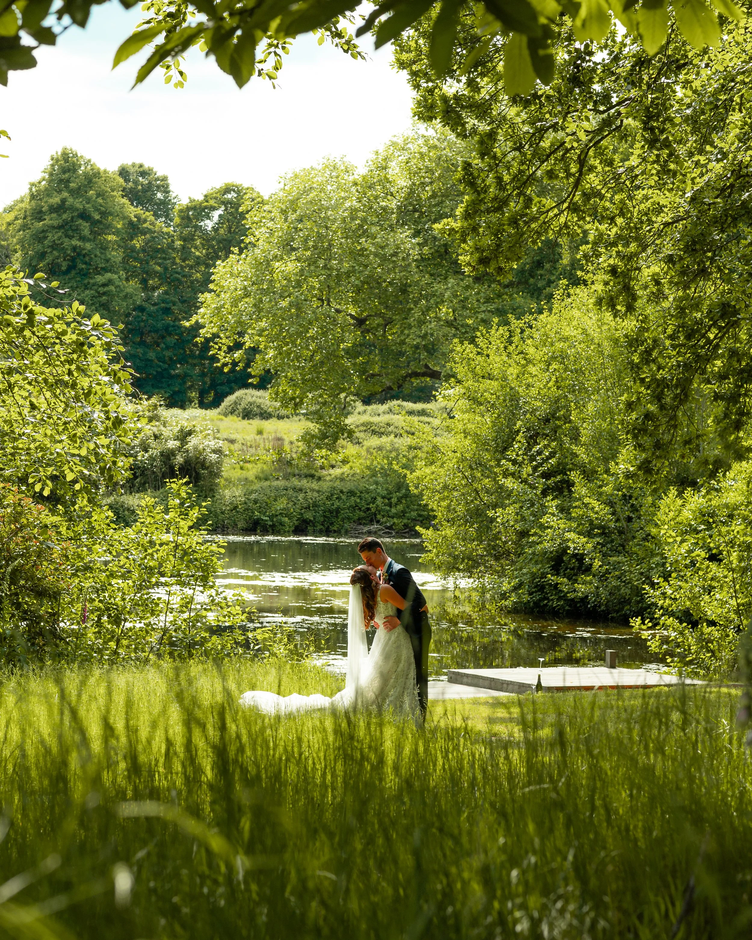 A newlywed couple kissing by a pond surrounded by trees and greenery on a sunny day.