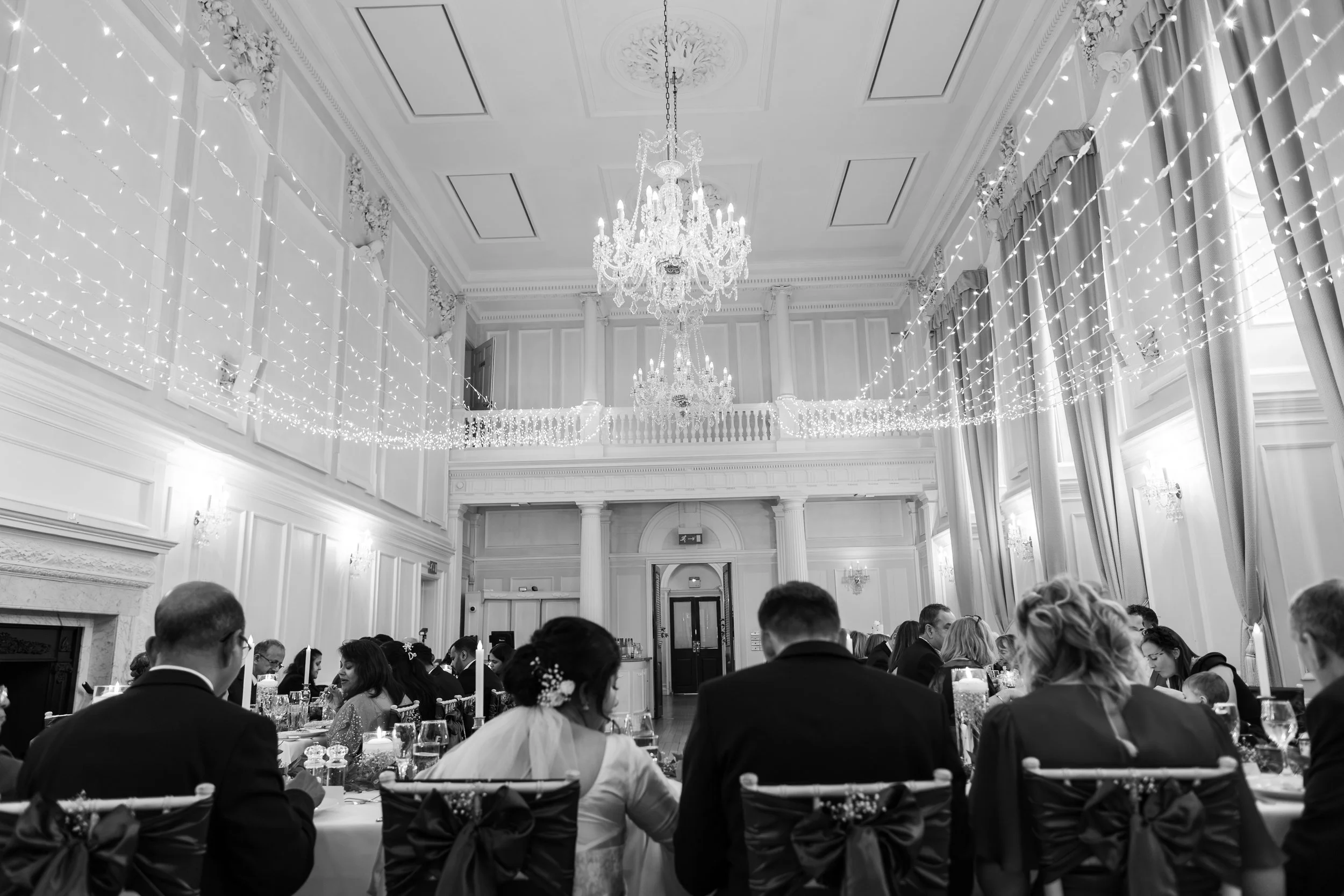 A black-and-white photo of a wedding reception in an elegant, ornate room with high ceilings, chandeliers, and string lights. People are seated at tables, with the bride and groom at the center of the image.