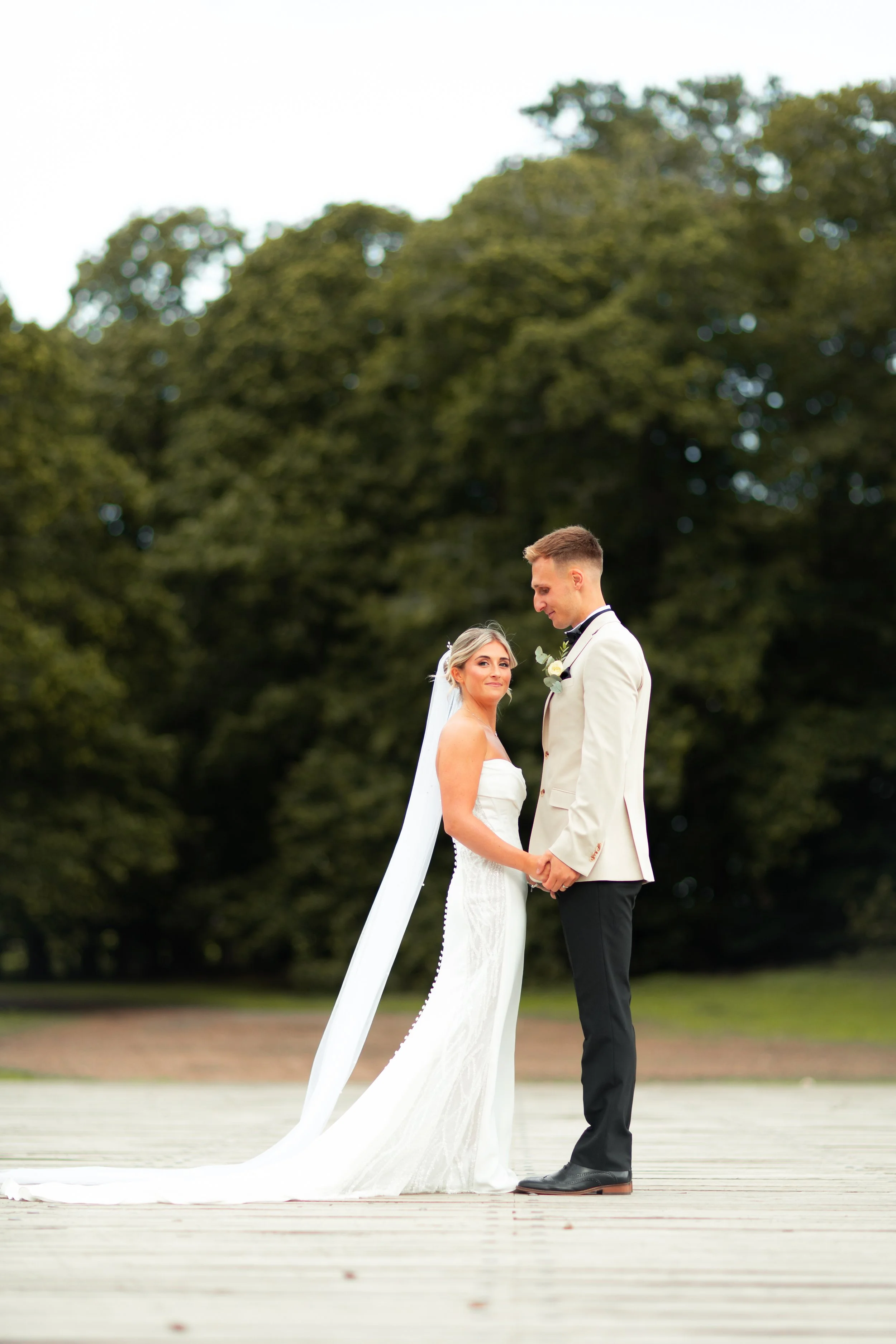 A bride and groom are holding hands and looking at each other during their outdoor wedding ceremony. The bride wears a white dress with a long train, and the groom wears a white tuxedo with black pants, both smiling at each other. The background feat
