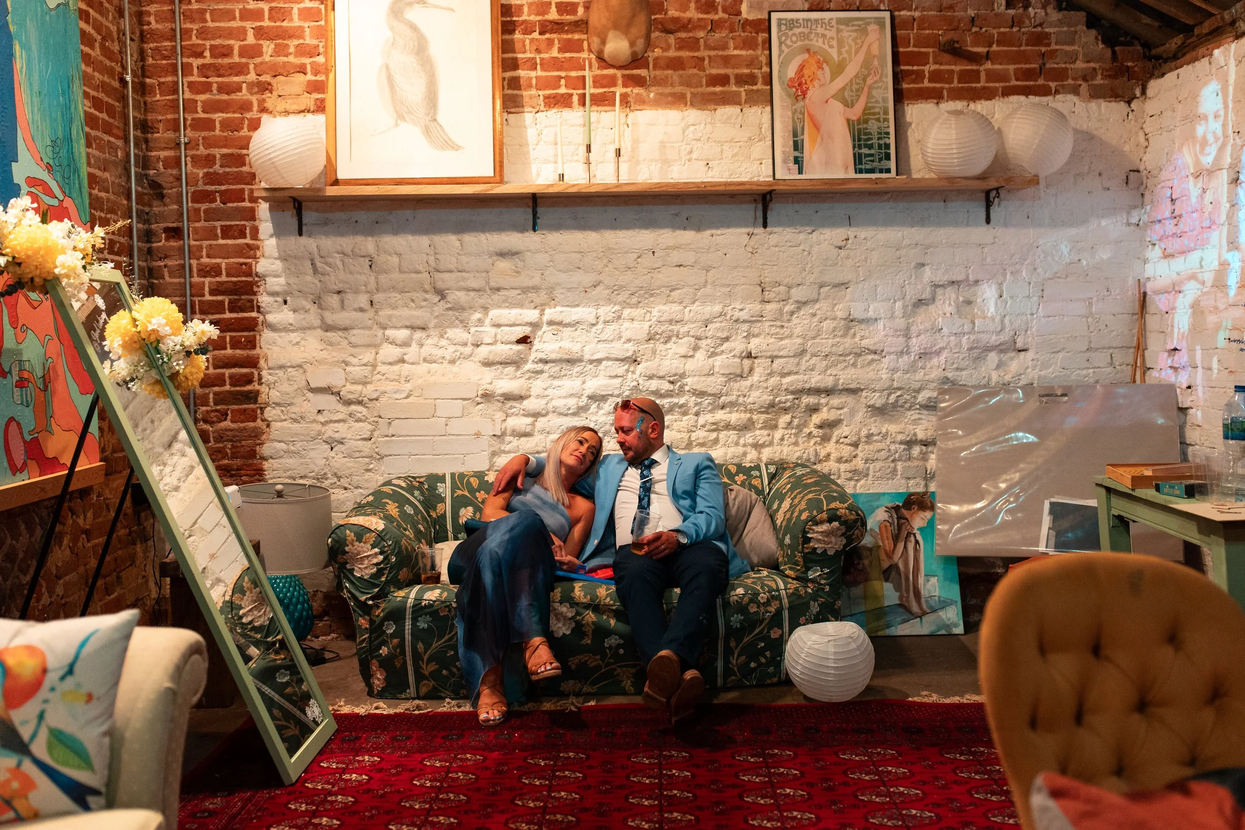 Two people sitting on a floral pattern couch, with one arm around each other, in a cozy, decorated indoor setting with exposed brick walls, artwork, and vintage decor.