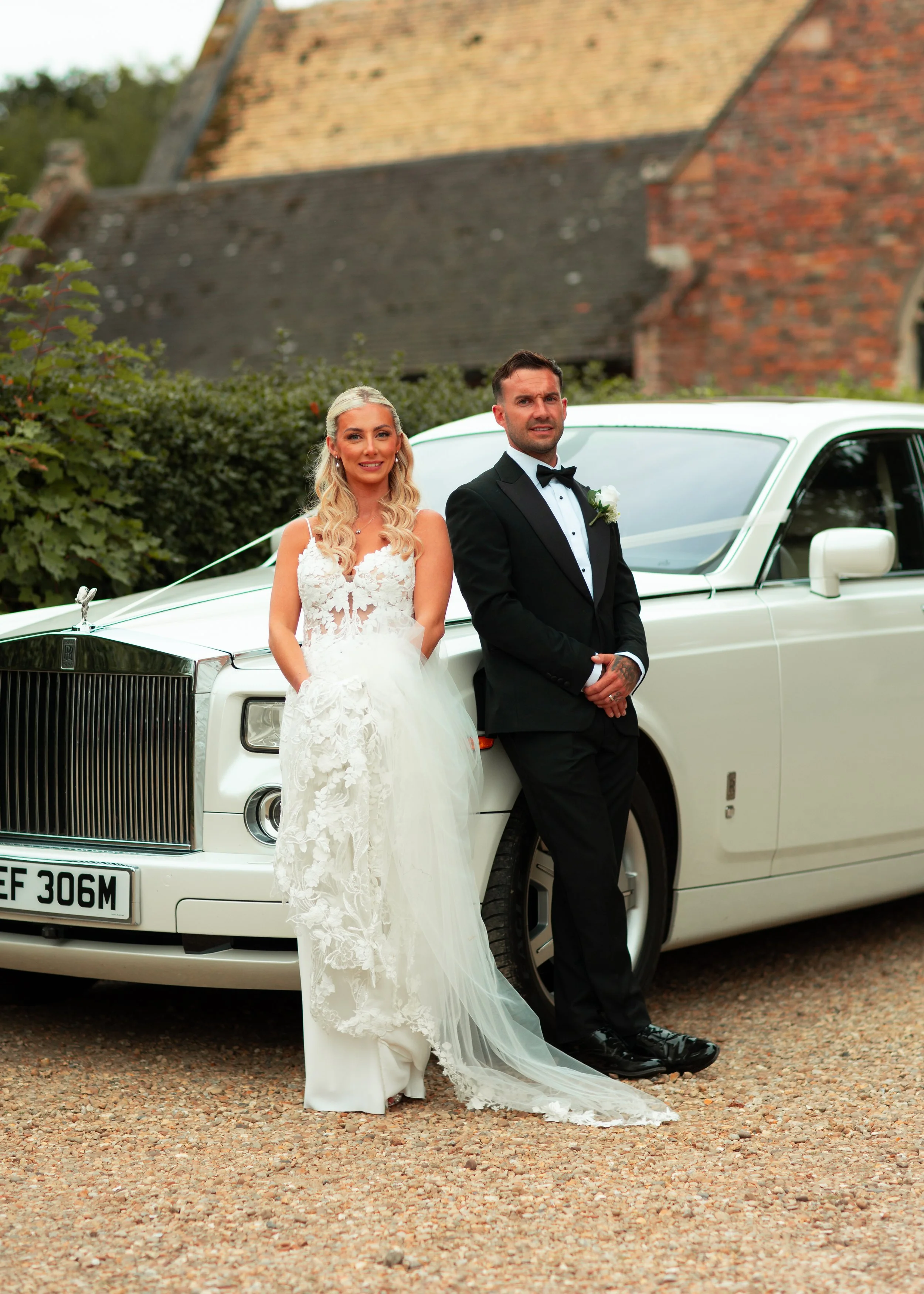 A bride and groom standing in front of a white limousine on their wedding day. The bride wears a lace wedding gown with a veil, and the groom wears a black tuxedo with a bow tie.