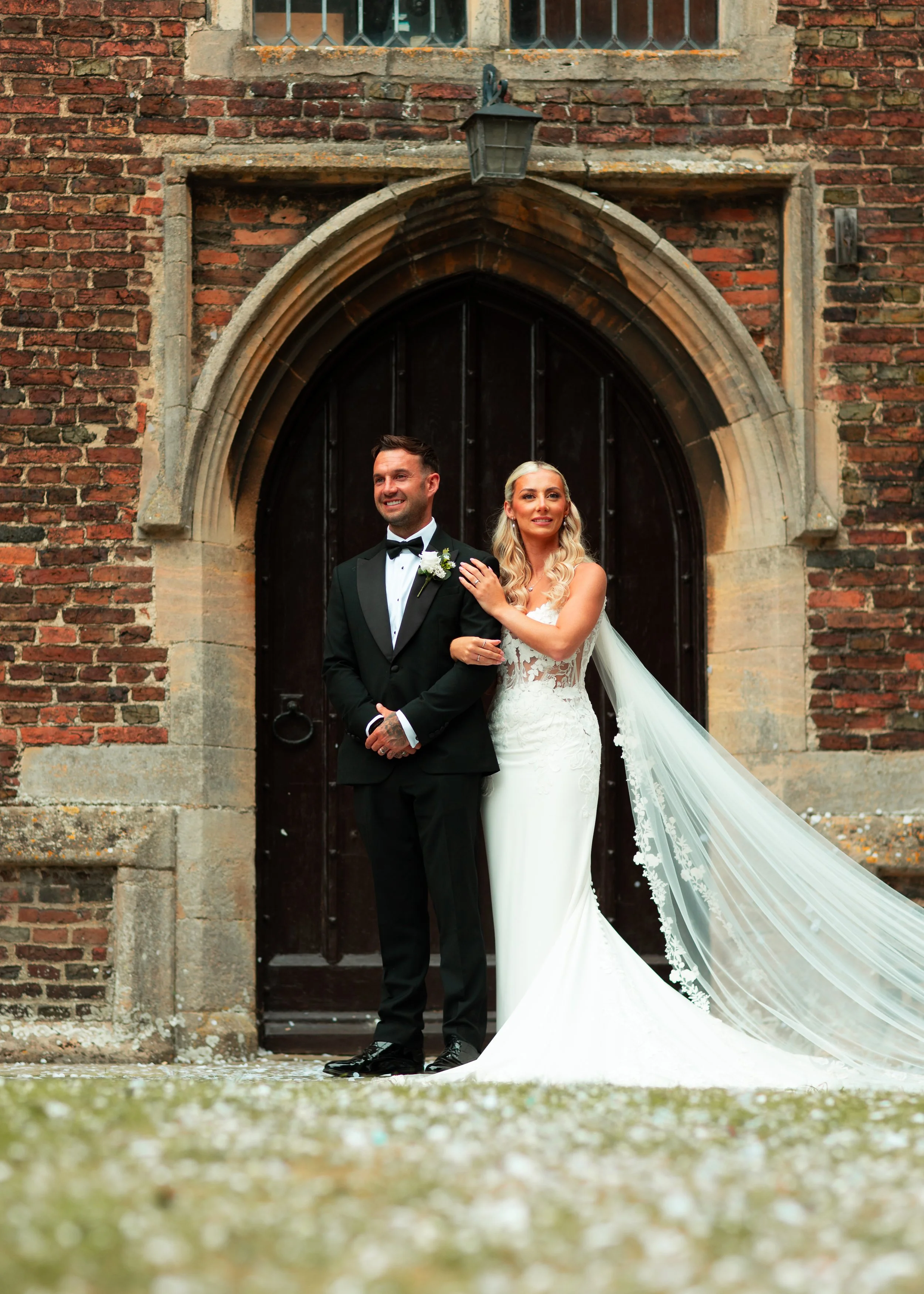 A bride and groom standing in front of a large wooden door with brick walls, the bride wearing a white lace wedding dress with a long train and veil, and the groom in a black tuxedo, both smiling.