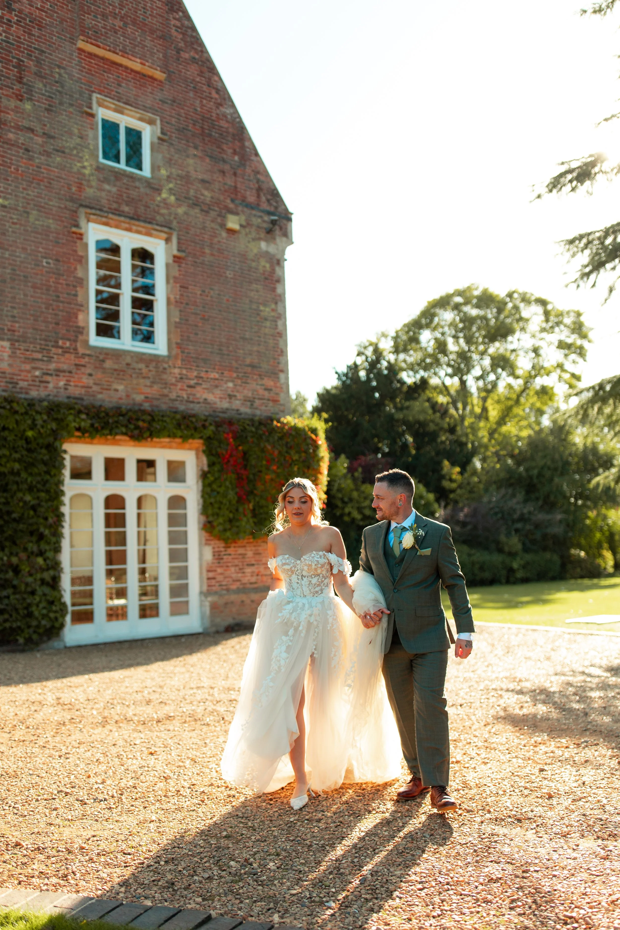 A bride in a wedding gown and a groom in a suit walk hand in hand outside a brick building during sunset.