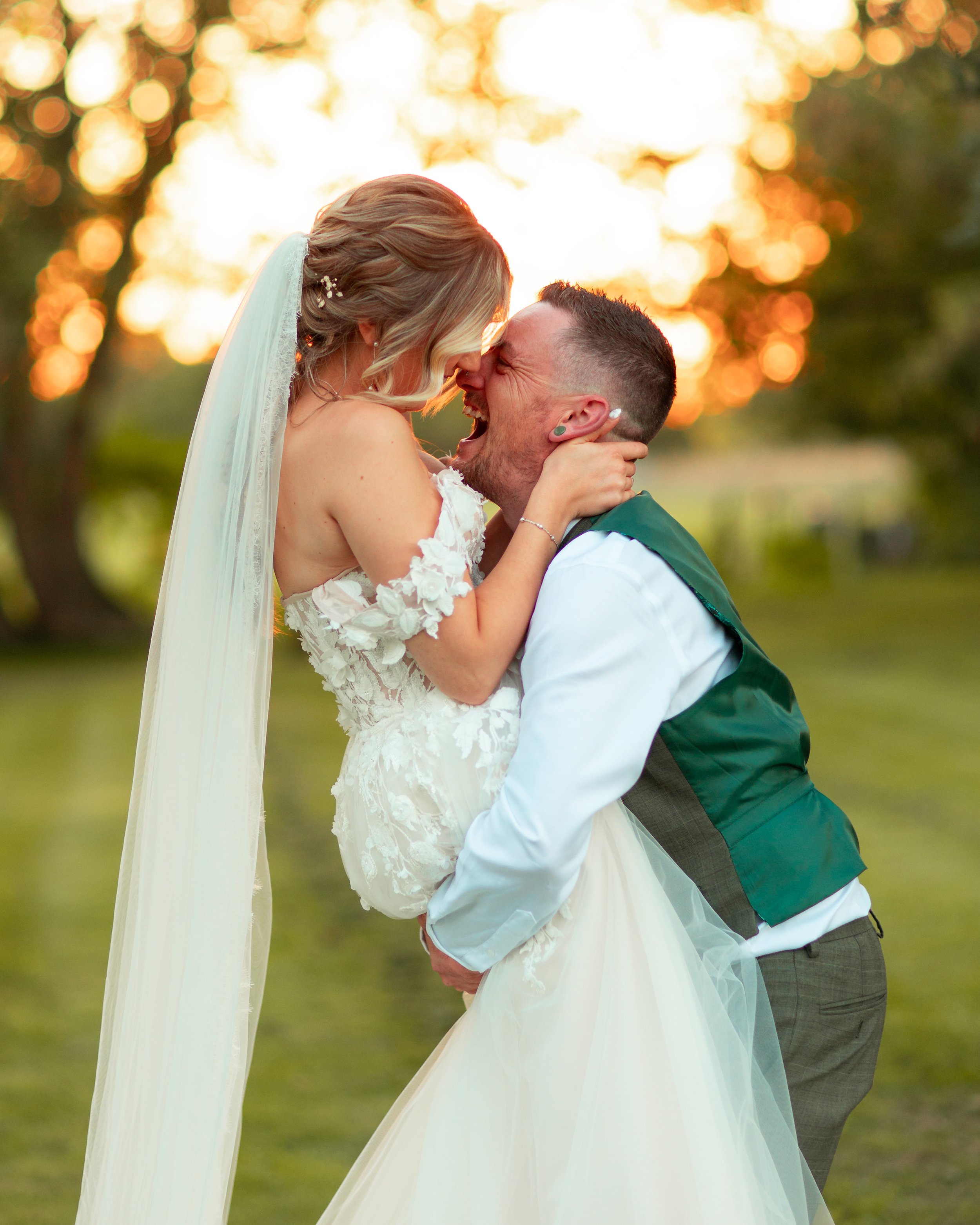 A bride and groom share a joyful moment outside during sunset, with the groom holding the bride in his arms as they smile and touch foreheads.