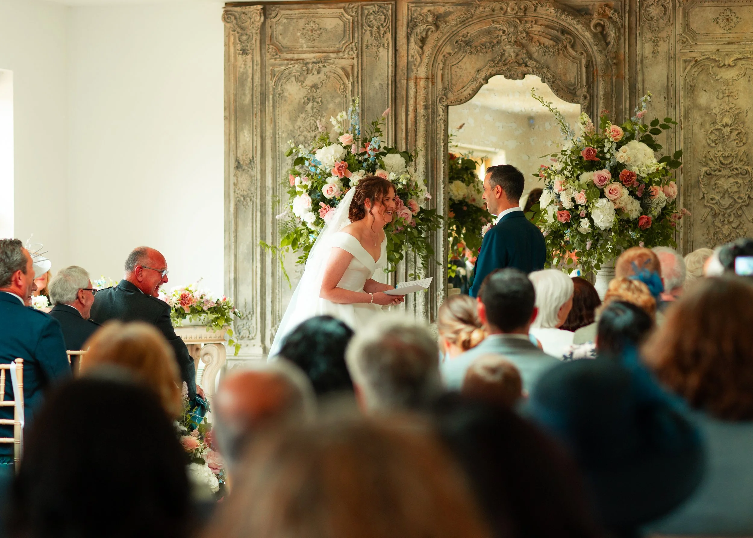Bride and groom exchanging vows during a wedding ceremony in a decorated indoor venue with large floral arrangements and ornate mirrors, surrounded by seated guests.