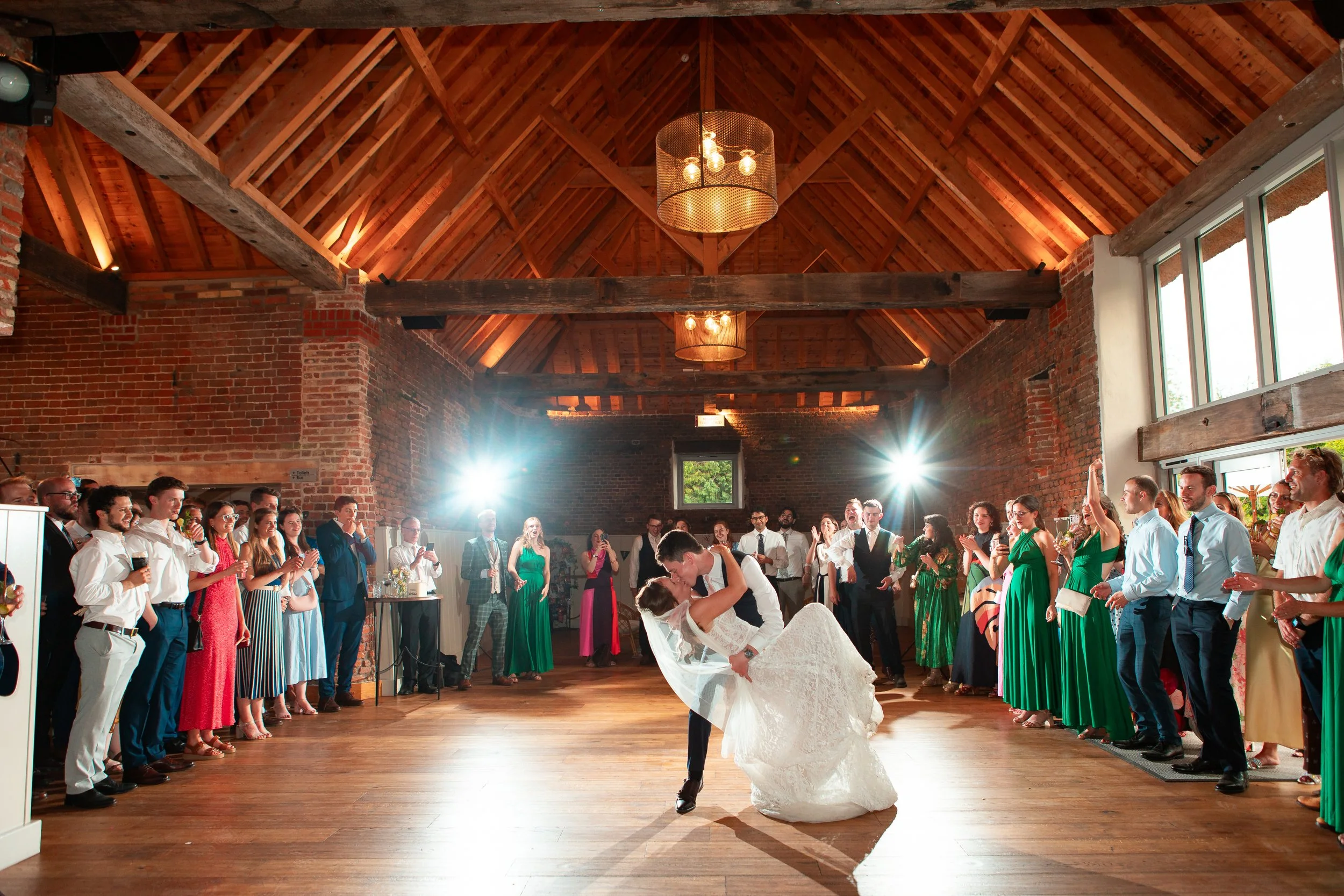 A bride and groom sharing a dance at their wedding reception, surrounded by guests in a rustic venue with wood beams and brick walls.