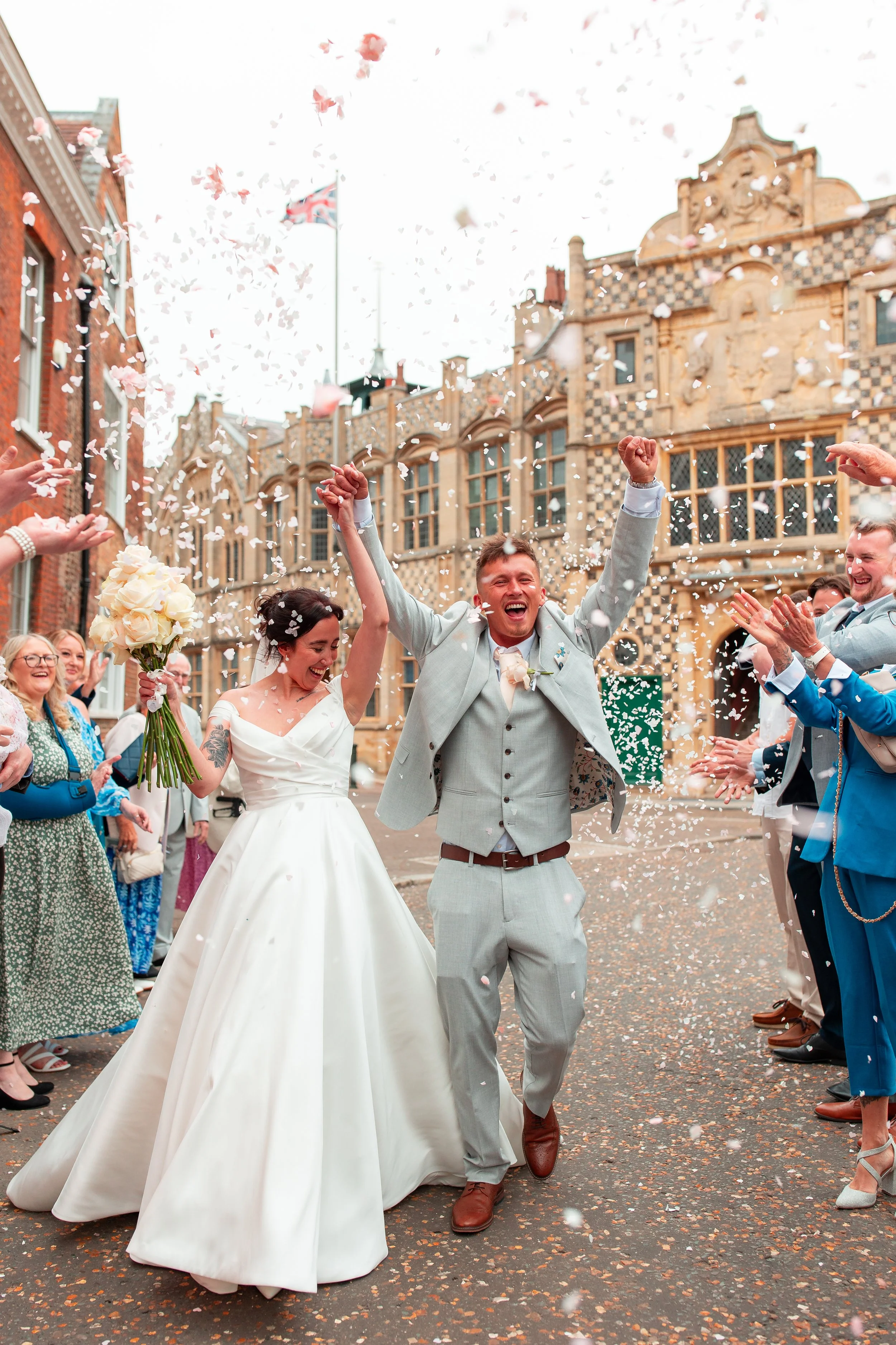 Joyful newlywed couple celebrating with friends and family outside on a cobblestone street, confetti flying all around, historic building in background.