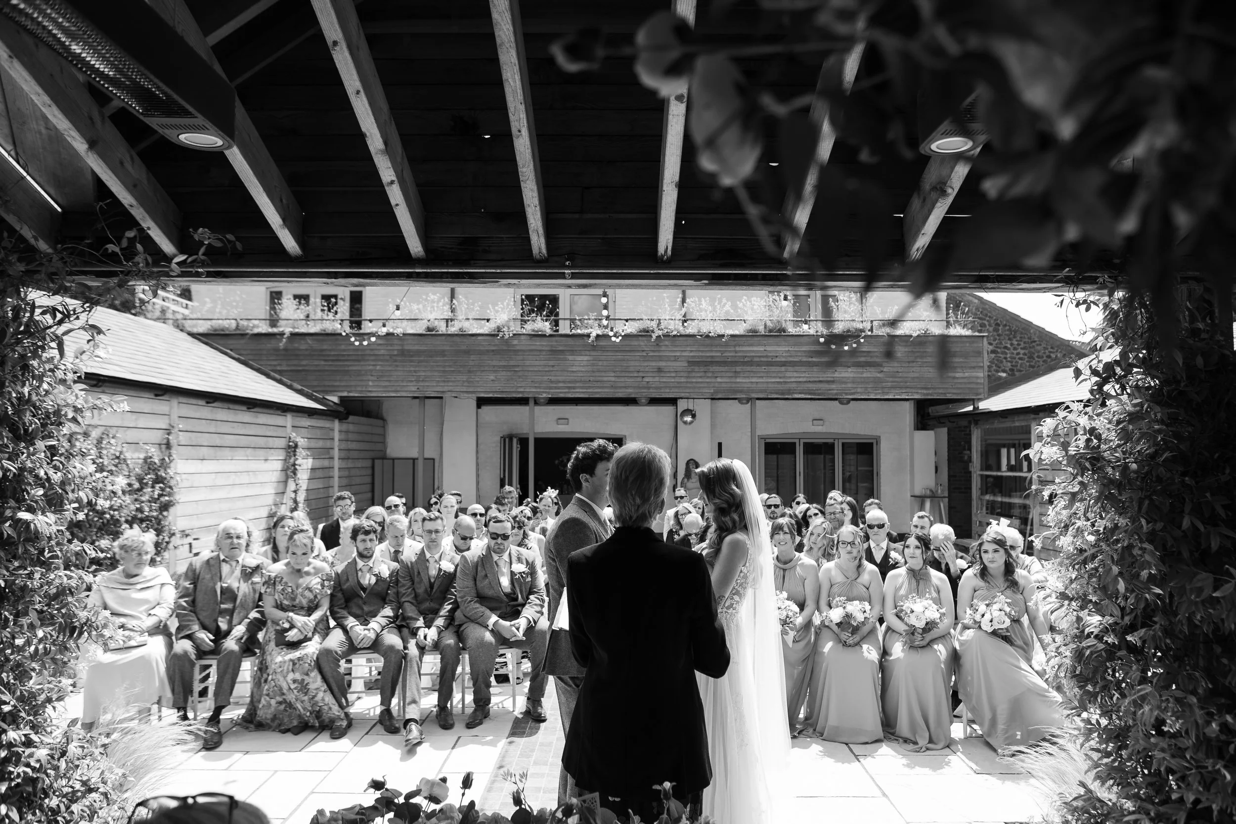 Black and white photo of a wedding ceremony with the bride and groom facing each other, surrounded by seated guests under a wooden outdoor structure.