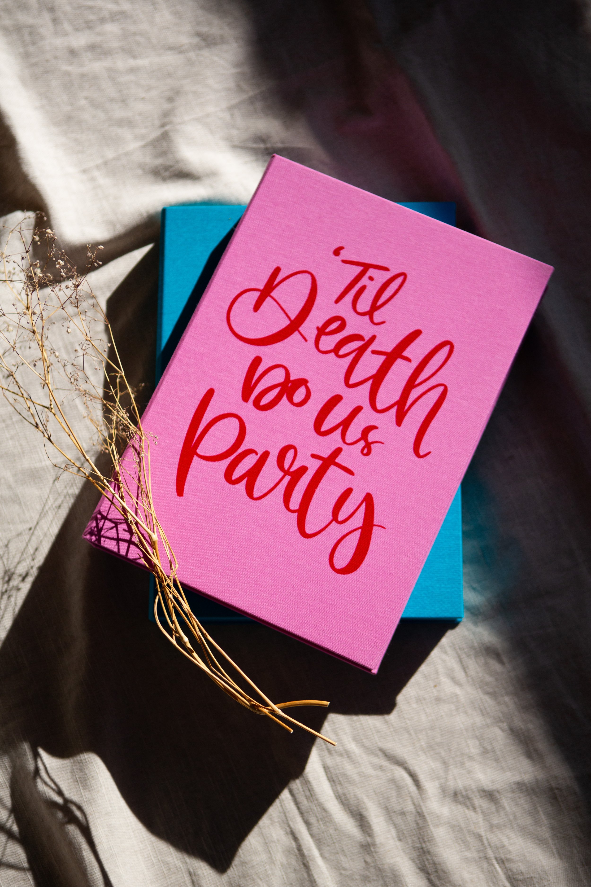 Pink greeting card with red text that says 'Til Death do us party', resting on a blue box with a dried plant beside it, on a light-colored fabric surface.
