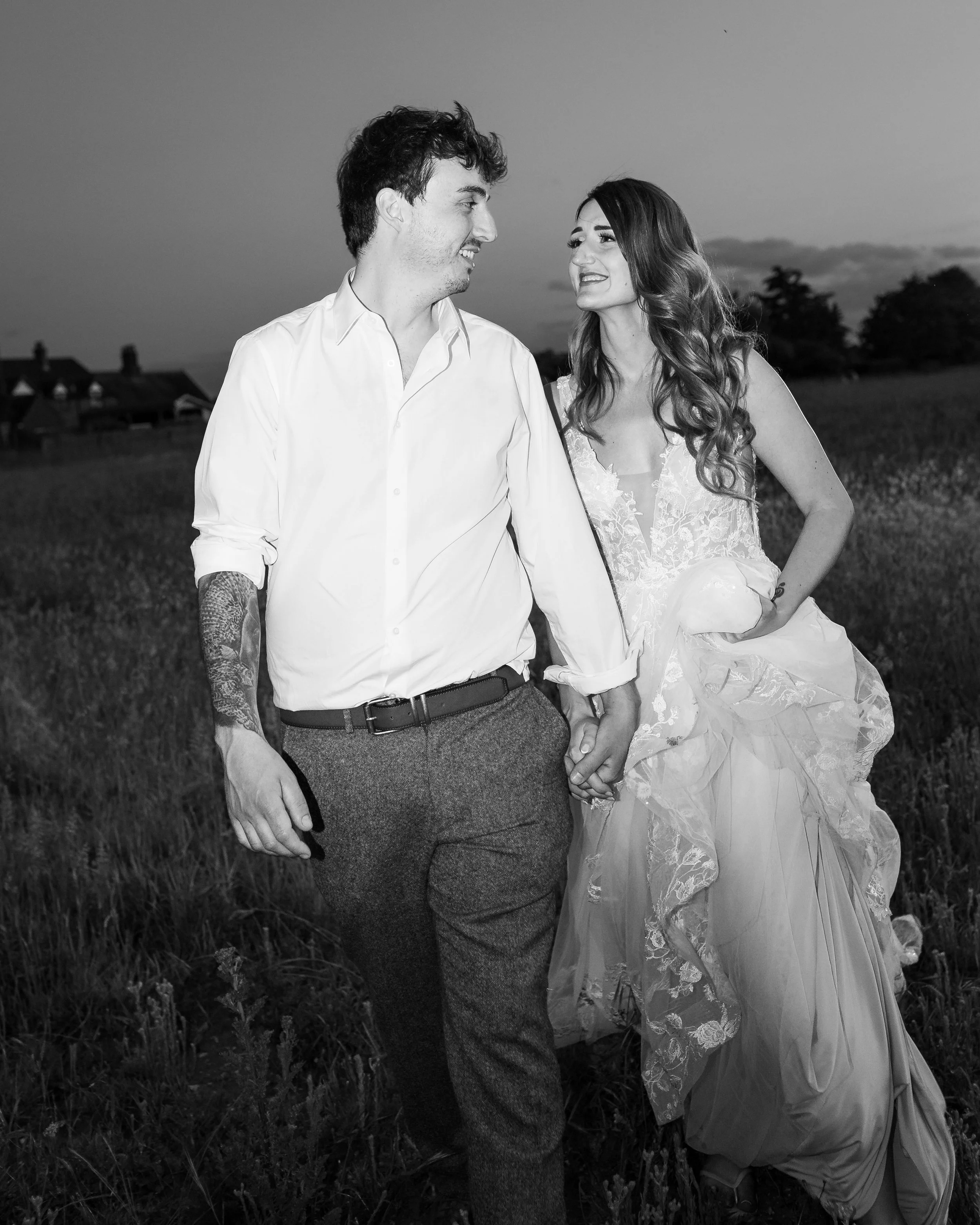 A couple walking hand in hand in a field, looking at each other and smiling, with houses in the background, in black and white.