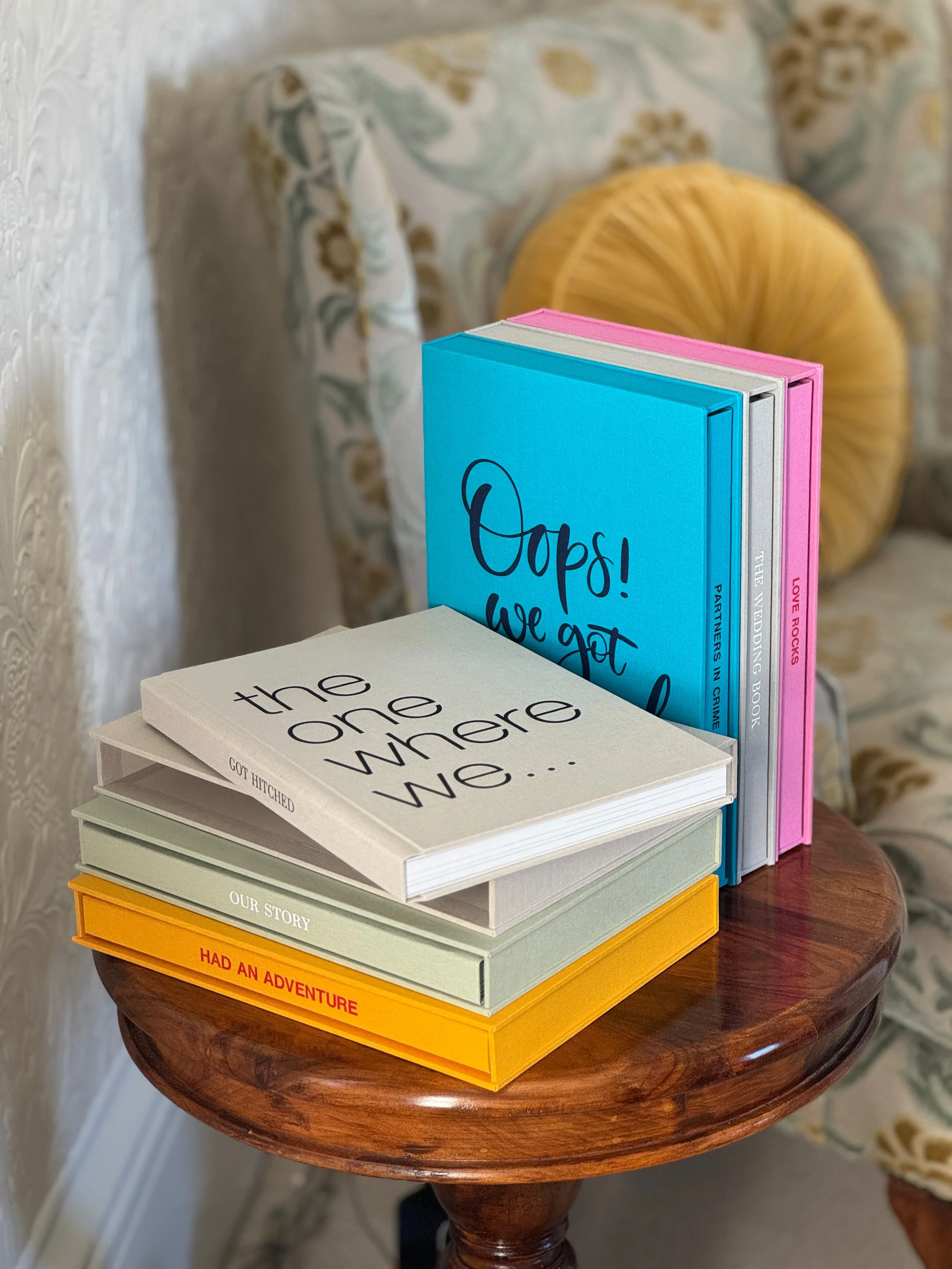 A stack of colorful books on a round wooden table with floral upholstery in the background.