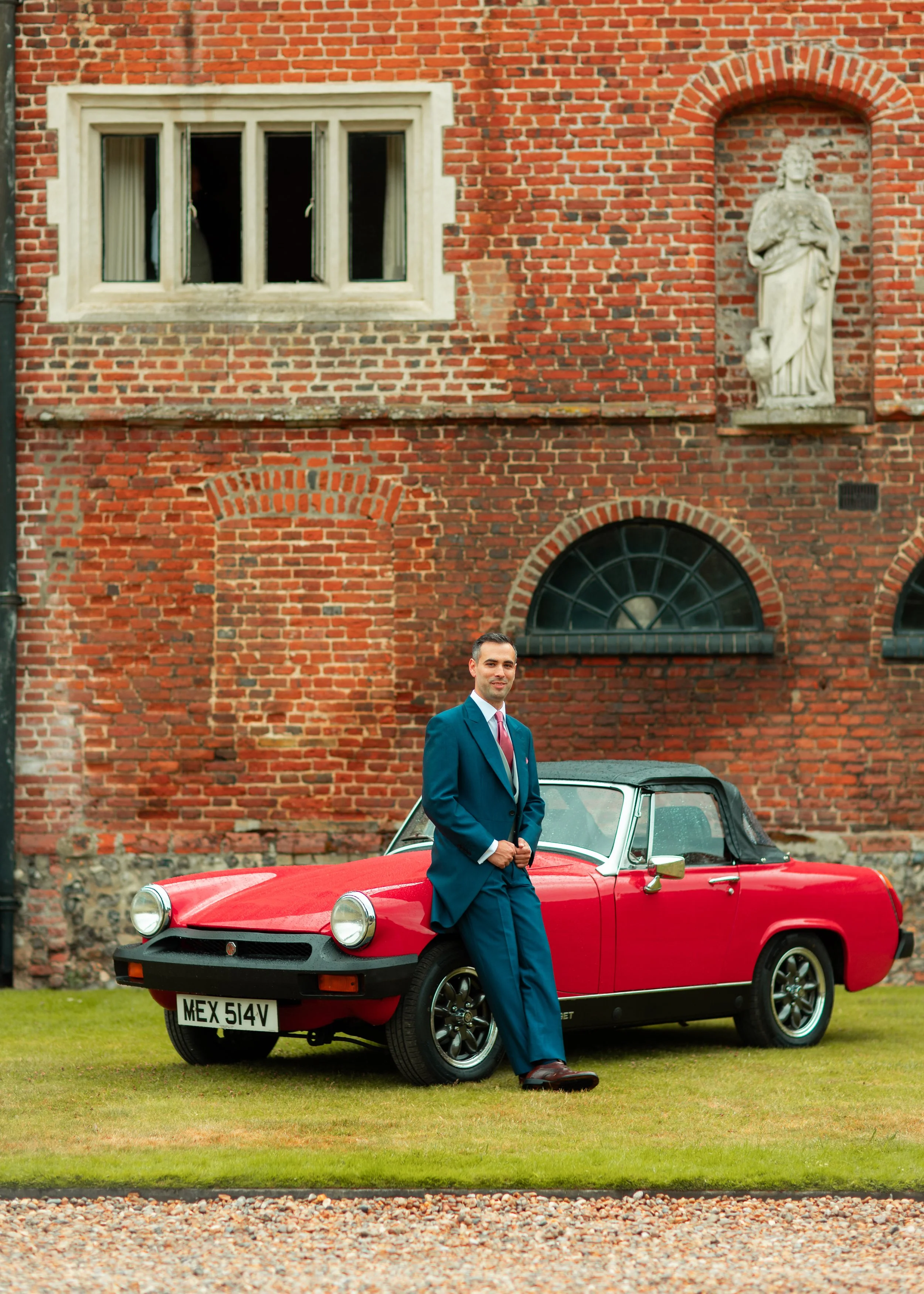A man in a blue suit stands beside a red convertible car with a black roof, in front of a brick building with a statue and arched windows.