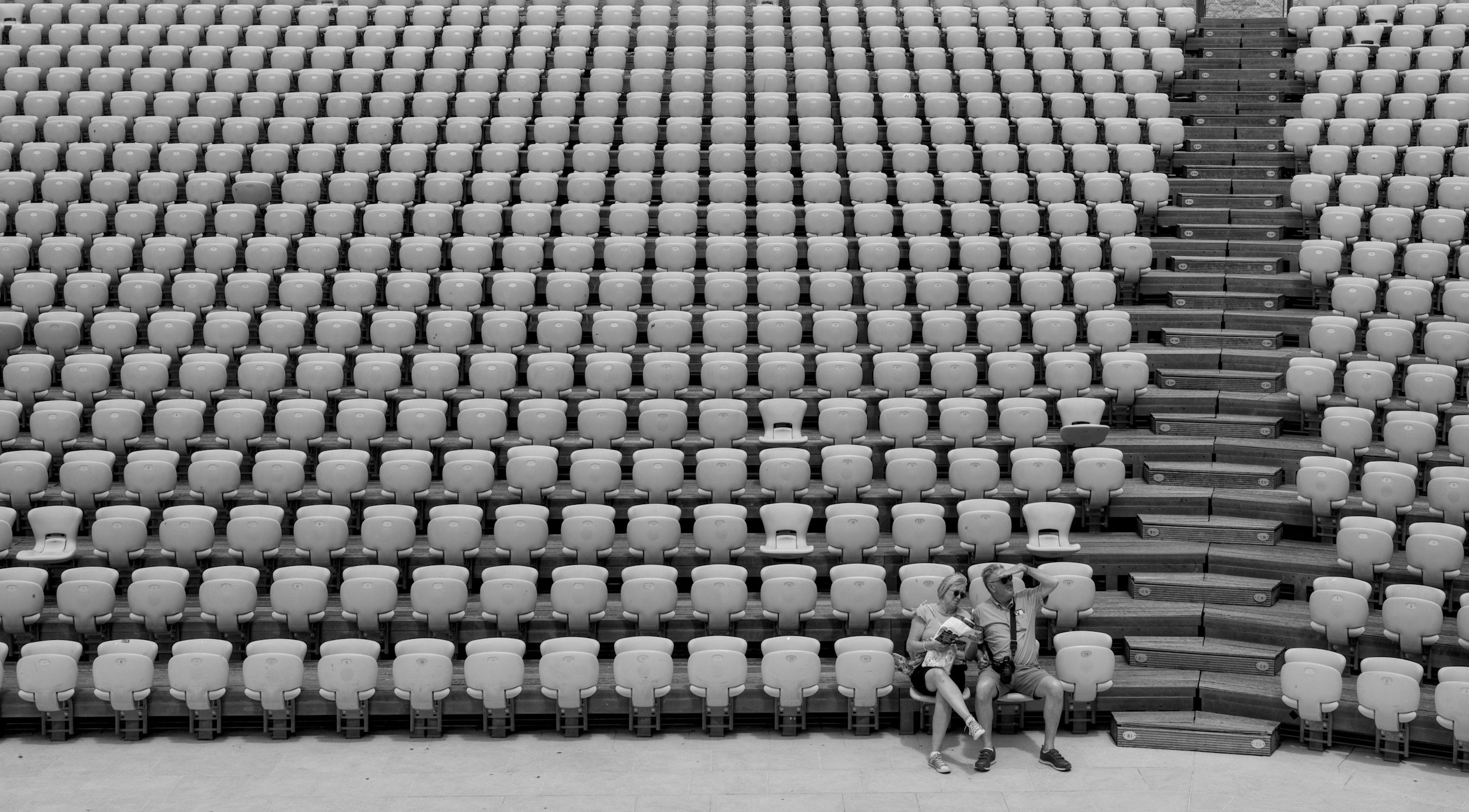 Two people sitting on a bench among rows of empty outdoor stadium seats, with some stairs in the background.