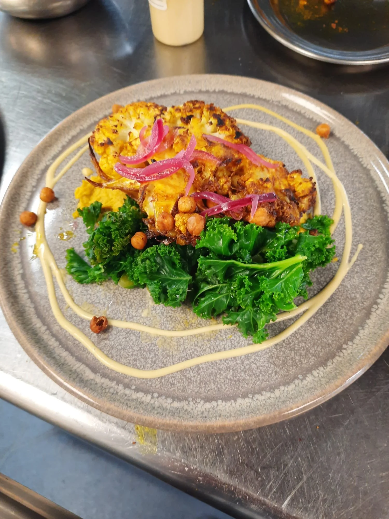 A plated dish of roasted cauliflower topped with pink pickled onions and crispy chickpeas, served with a side of curly kale and a decorative drizzle of sauce on the plate.