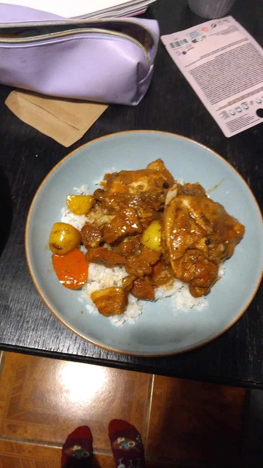 Plate of rice topped with chicken curry containing potatoes and carrots, on a dark table with a receipt, purple purse, and tan napkin nearby.