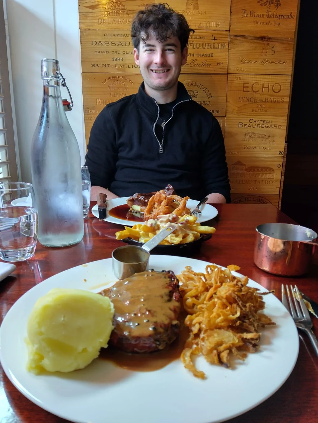 A smiling man sitting at a restaurant table with a plate of mashed potatoes, a piece of meat with gravy, and fried onions in the foreground. There are also plates of food on the table, a water bottle, and glasses of water. The background features a wooden wall with etched wine and winery names.