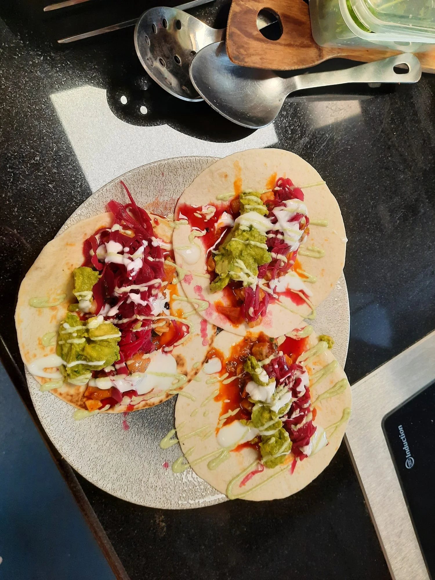 Three tacos on a white plate with shredded red cabbage, green guacamole, drizzled sauces, and chopped vegetables, placed on a black countertop.