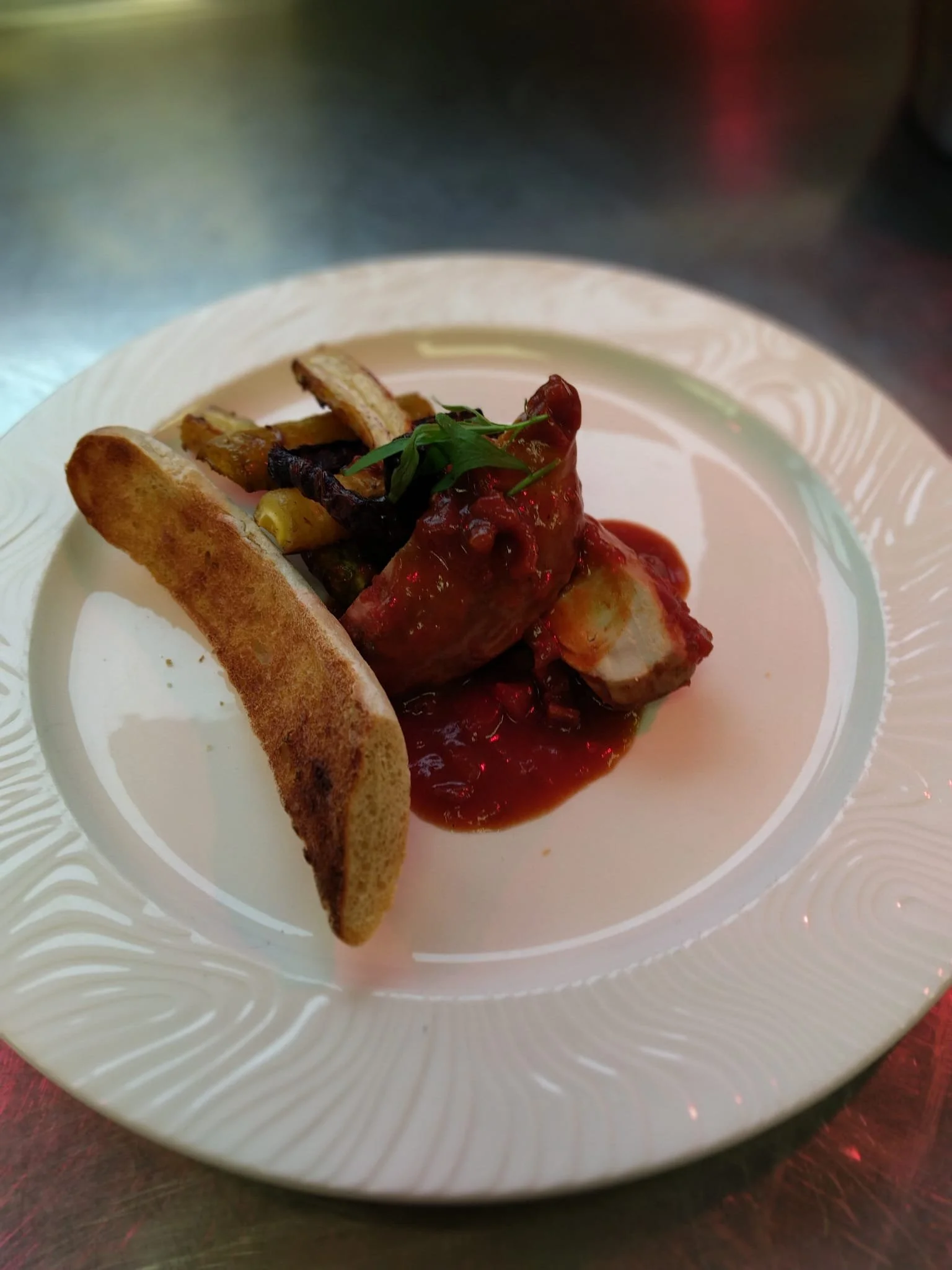 Plate of food with a piece of meat covered in red sauce, grilled vegetables, and a slice of toasted bread, garnished with a green herb.