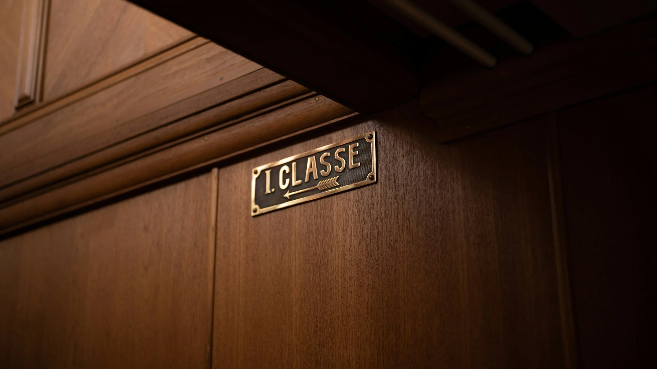 Close-up of a wooden door with a brass nameplate that reads '1. CLASSE' and an arrow pointing to the left.