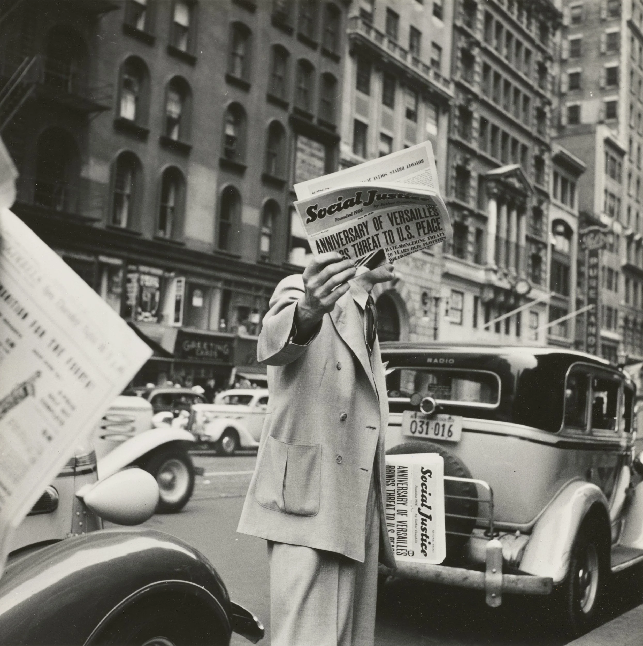 A man in a light-colored suit holds up a newspaper with a headline about the anniversary of Versailles and a threat to U.S. peace, standing on a busy city street with cars and tall buildings in the background.