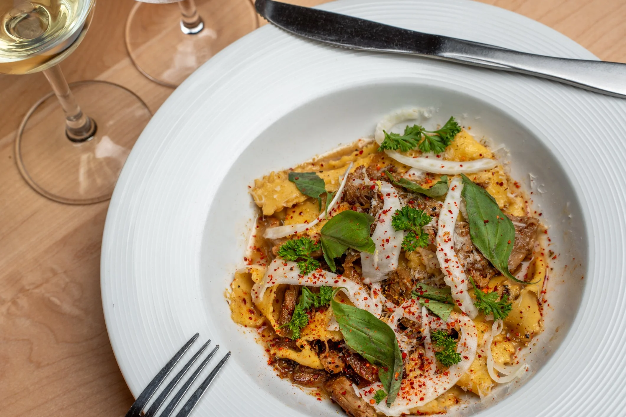 Plate of pasta with herbs, vegetables, and seasoning, served with a fork and knife, on a wooden table with glasses of white wine.
