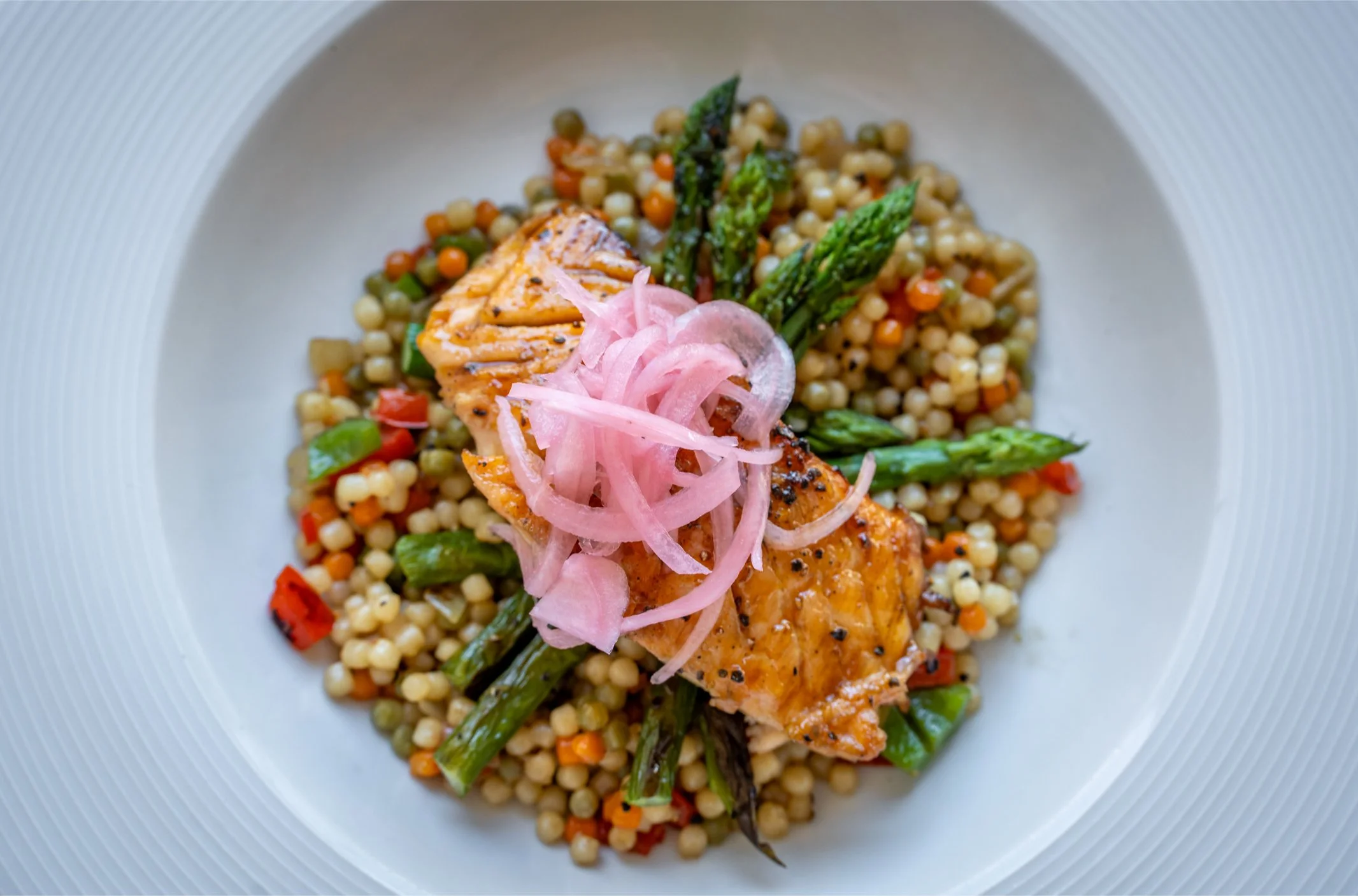 A plate of grilled salmon served over a bed of Israeli couscous with vegetables, garnished with asparagus, red bell peppers, and pickled onions.