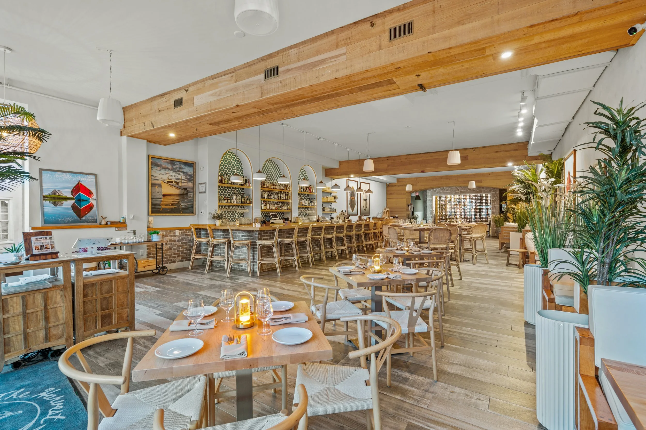 A cozy seafood restaurant with wooden floors and walls, set with tables, chairs, and a bar area with shelves of bottles. Decor includes nautical artwork, potted plants, and ambient lighting.