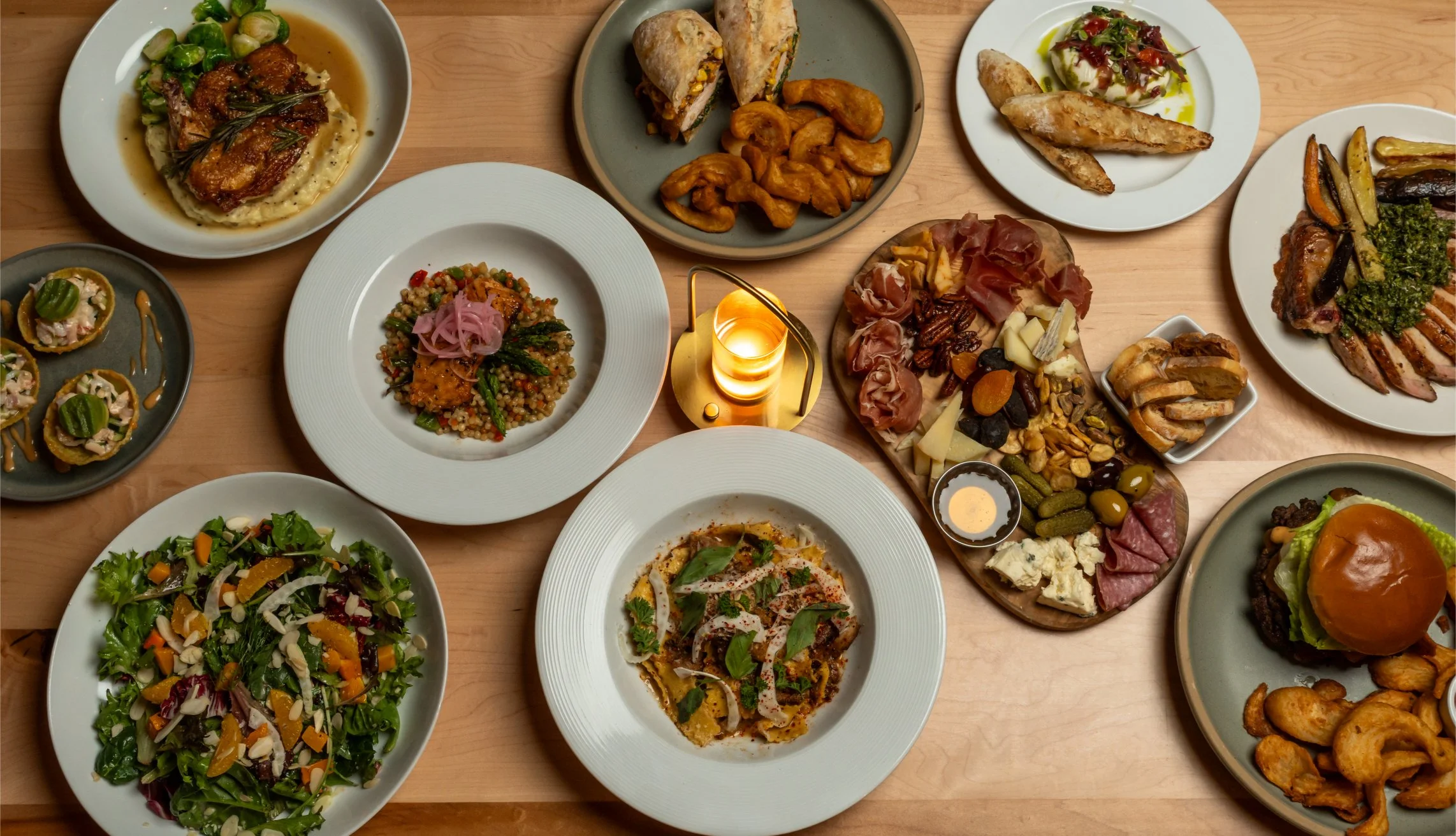 A variety of dishes on a wooden table, including salads, grilled meats, cheese board, wraps, and fried appetizers.