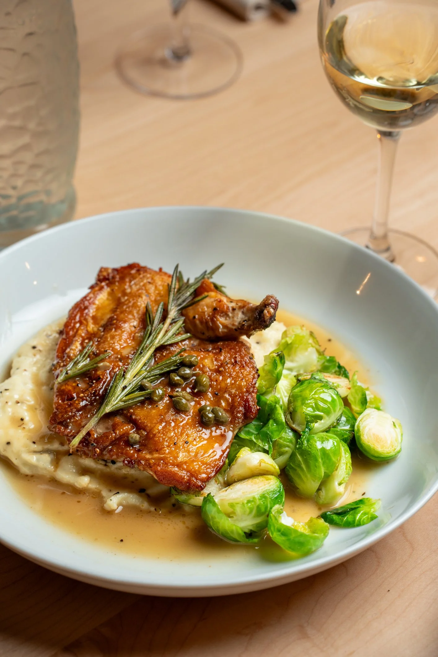 A plate of food featuring a roasted chicken leg garnished with rosemary and peppercorns, served with mashed potatoes and Brussels sprouts. A glass of white wine is in the background on a wooden table.