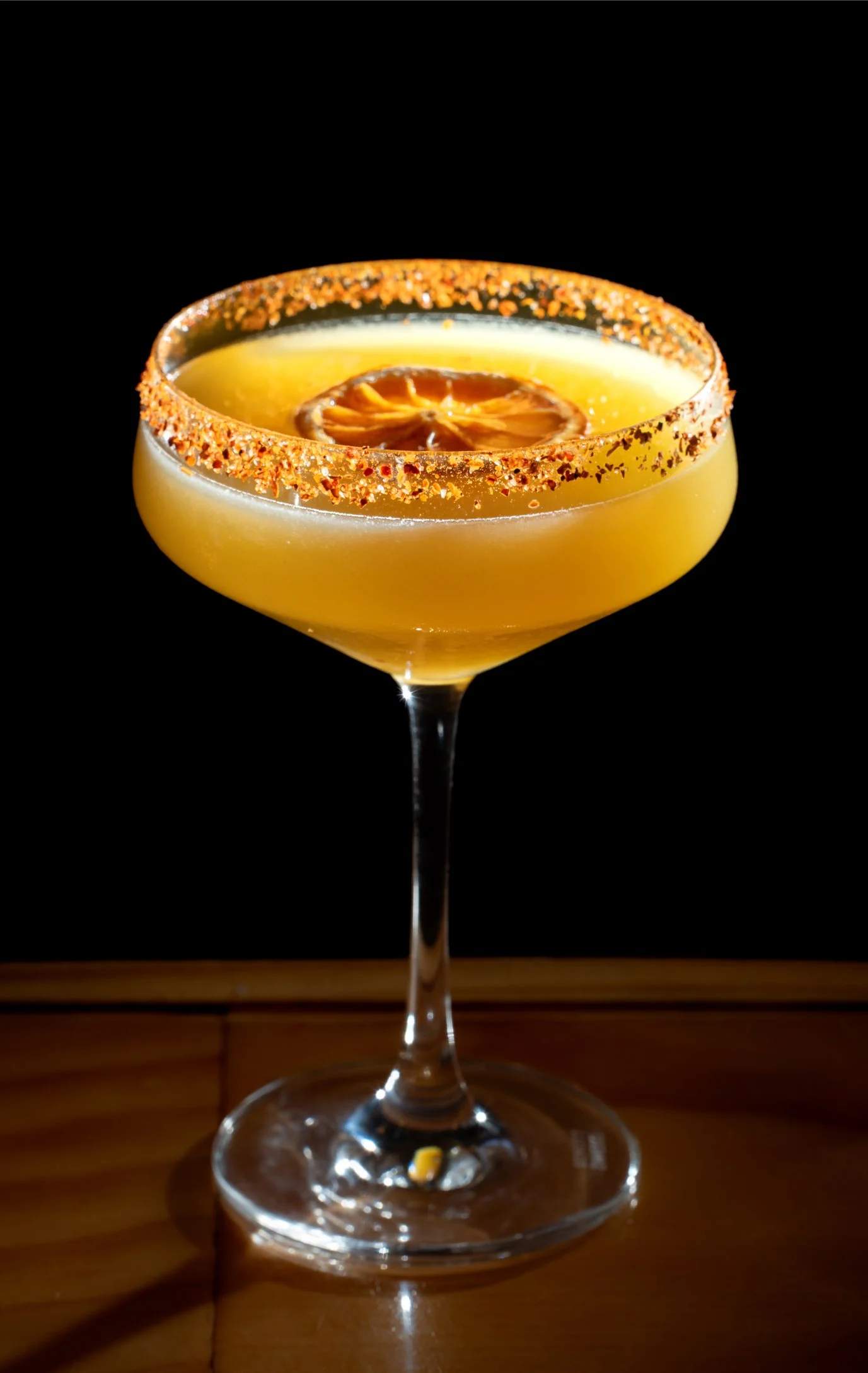 Yellow cocktail in a coupe glass with a salted rim and a dried lemon slice garnish on top, placed on a wooden surface against a black background.