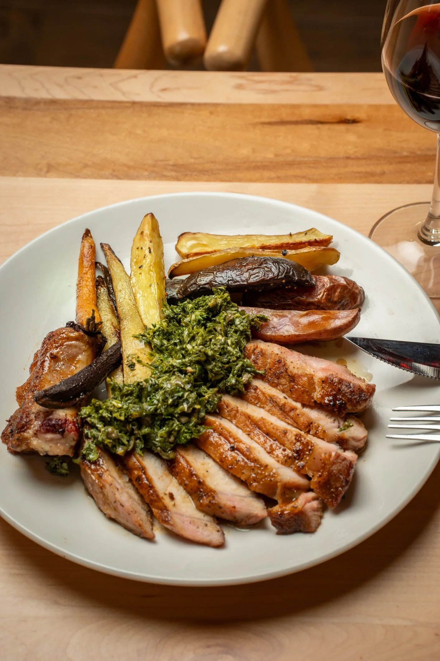 A plate of cooked sliced chicken breast topped with herbs, alongside roasted vegetables including potato wedges, carrots, eggplant, and peppers, with a glass of red wine on a wooden table.