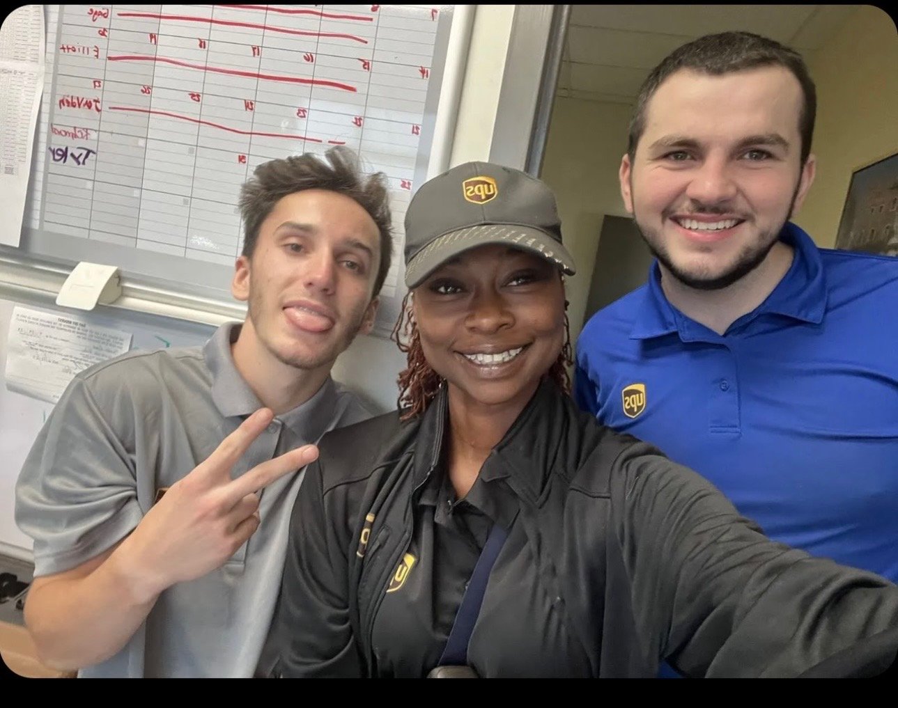 Three UPS employees taking a selfie inside a UPS store, with a whiteboard containing scheduling information in the background.