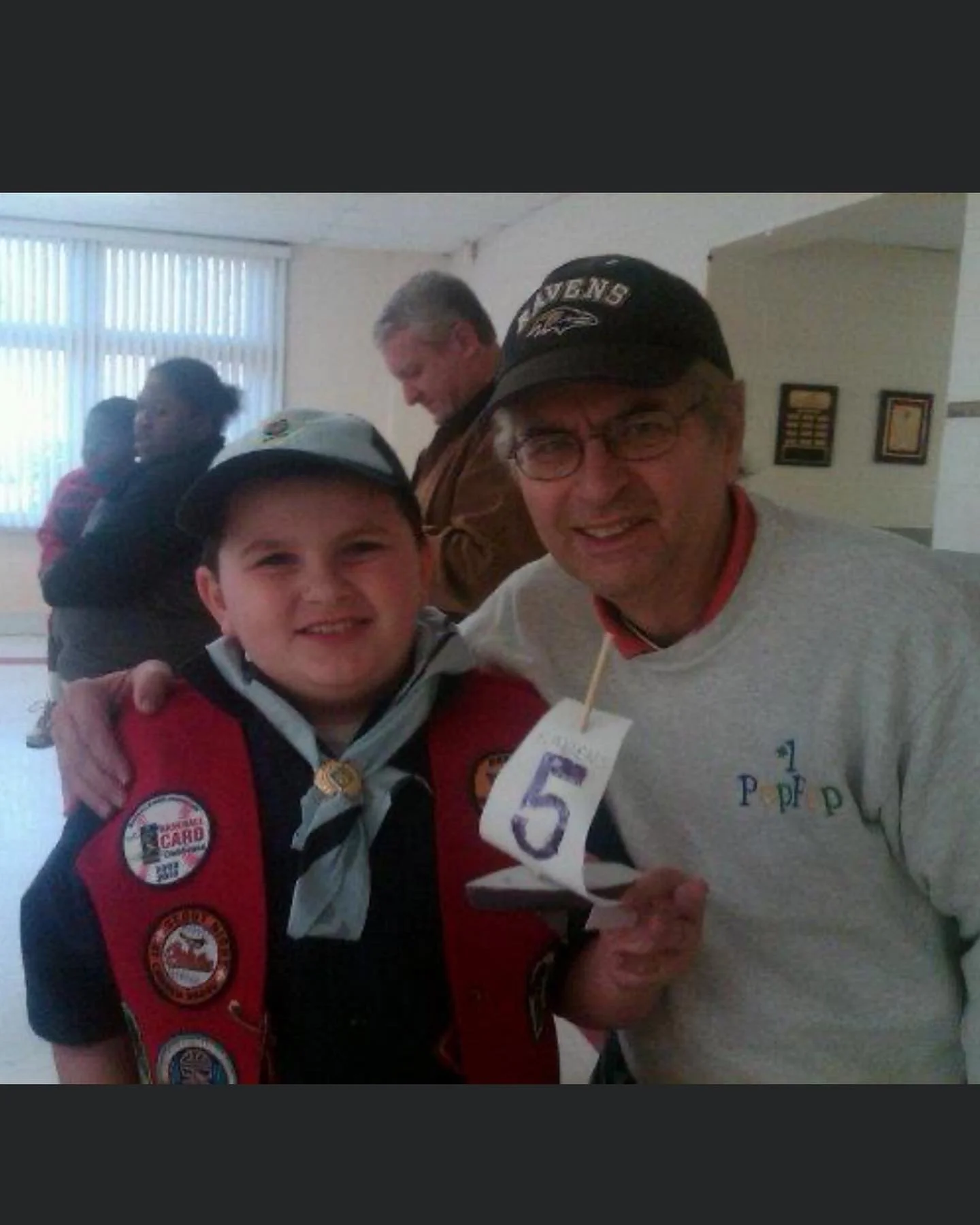 A young boy in a Cub Scout uniform standing next to an older man wearing a cap and glasses. They are indoors, smiling at the camera, with the boy holding a paper tag with the number five. Other people are visible in the background.