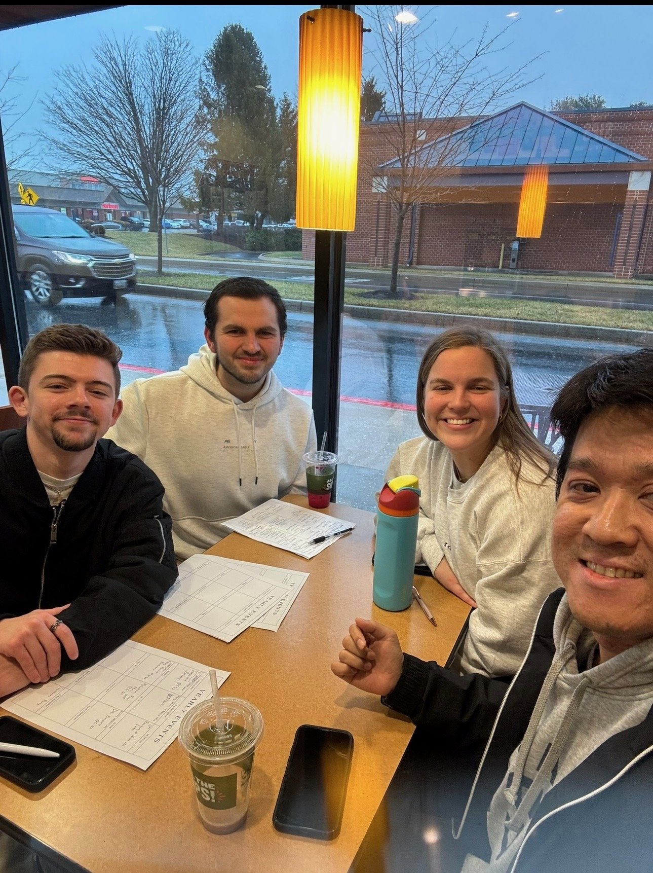 Five people sitting at a restaurant table with menus, drinks, and a water bottle, smiling at the camera during a rainy day outside.