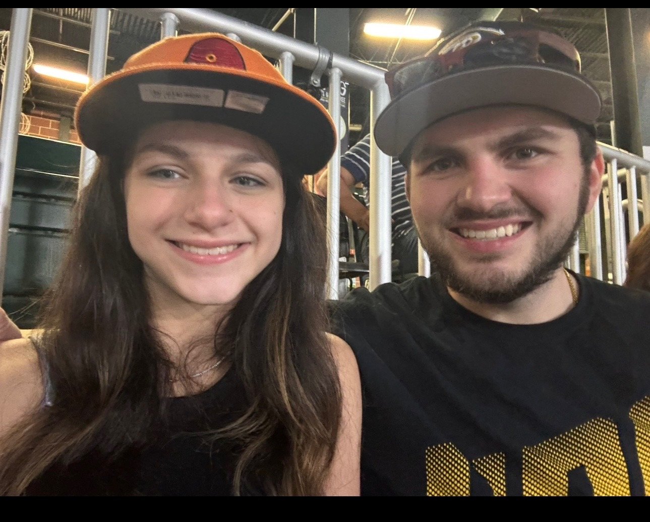A smiling young woman and man taking a selfie together, both wearing baseball caps, in a casual indoor setting with metal railings in the background.