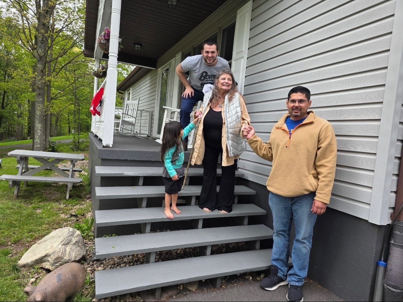 A group of five people, including two children, standing on the stairs of a house, smiling and holding hands. Two of them are on the porch and three are on the stairs. The house has gray siding and a porch with hanging baskets and a Canadian flag. The surrounding area includes trees, grass, a picnic table, and decorative rocks.