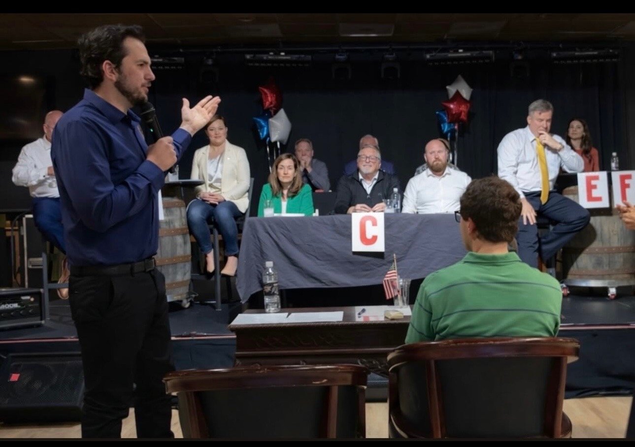 A man in a blue shirt holding a microphone and speaking to an audience at a panel discussion with seven other panelists sitting at a table on a stage decorated with red, white, and blue balloons. The audience member visible in the foreground is sitting in a green striped shirt.
