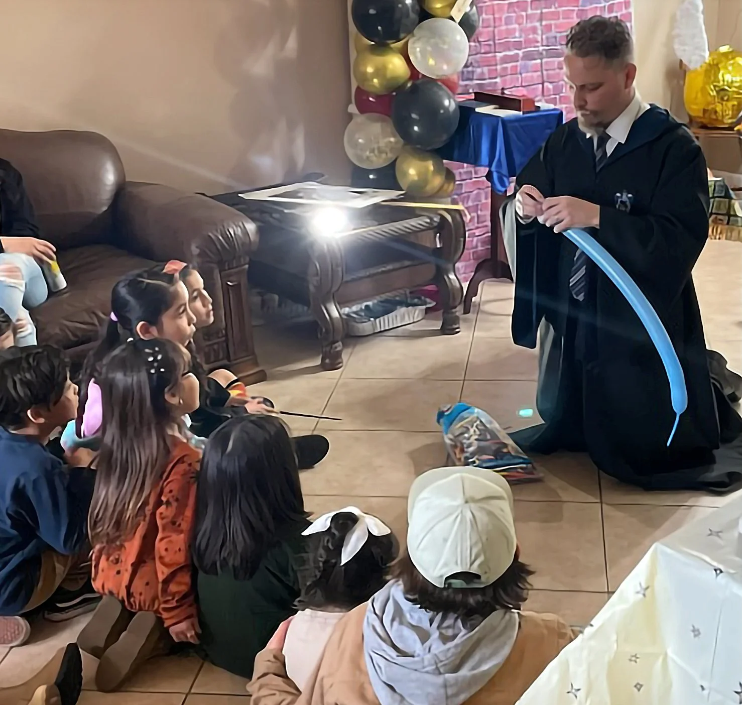A man dressed in black with a blue light is kneeling on the floor performing a magic trick for a group of children sitting on the floor. The children are attentively watching him, some with bunny ear headbands. There are balloons and decorations in t