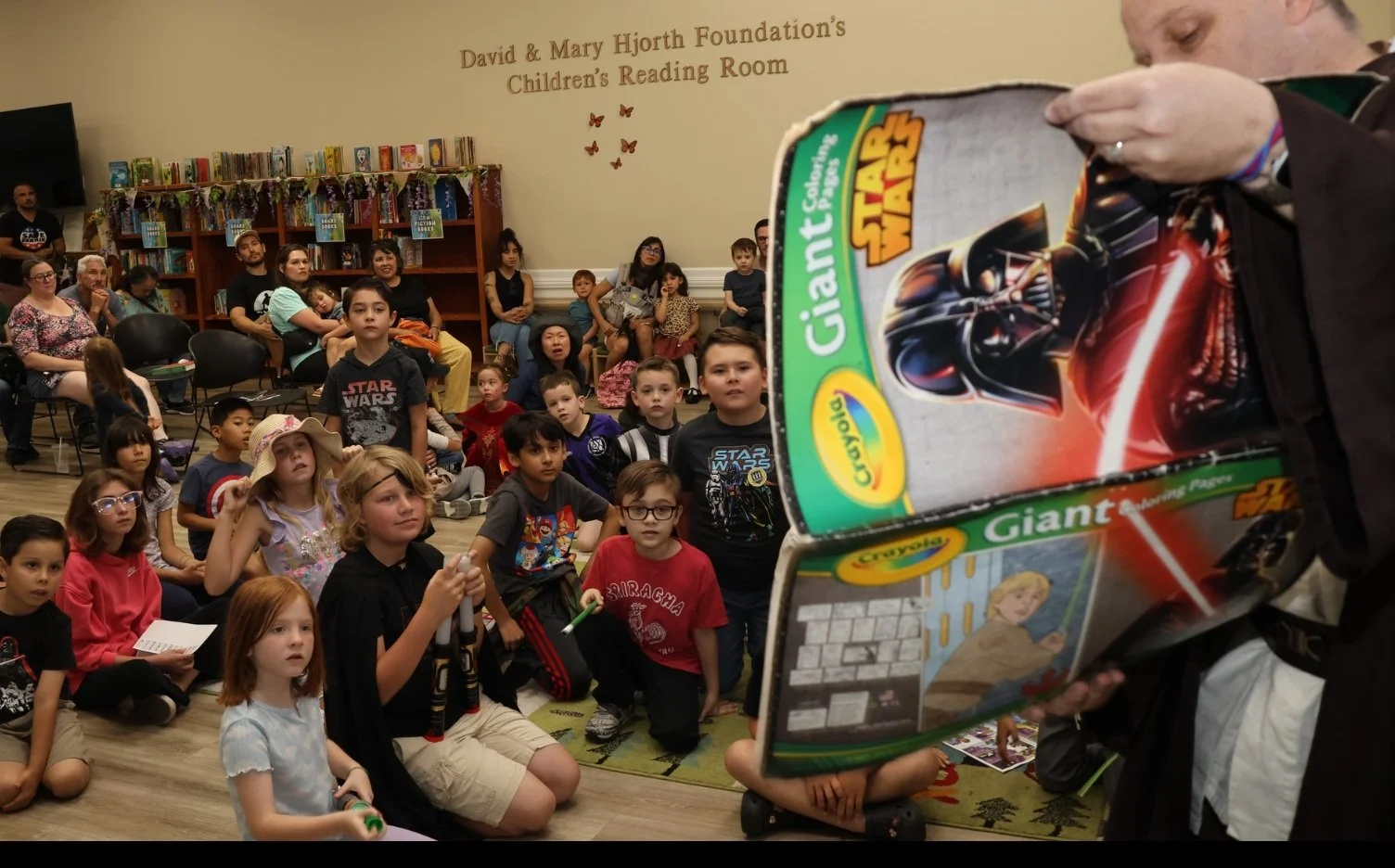 Children sitting on the floor and adults seated in chairs inside a children’s reading room, listening to a person reading a Star Wars-themed story from a large Crayola Giant Coloring Pages book. The wall behind them has bookshelves and a sign that reads 'David & Mary Hjorth Foundation's Children's Reading Room.'