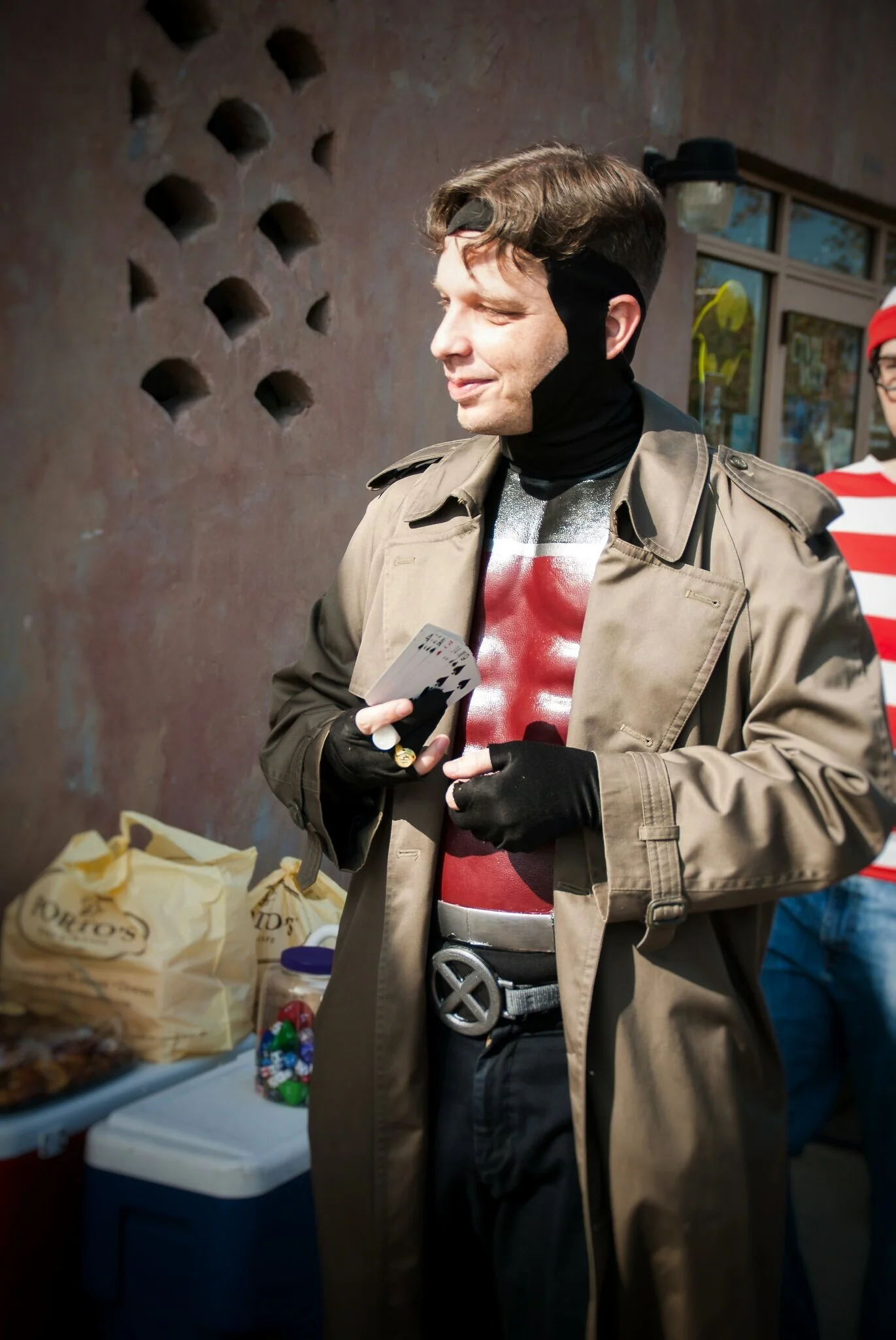 A man dressed as Colonel Sanders from KFC, with a trench coat, black gloves, and a badge, holding a hand of playing cards, standing outdoors near a table with snacks and a jar of candy, with a person dressed as Waldo from 'Where's Waldo?' in the back